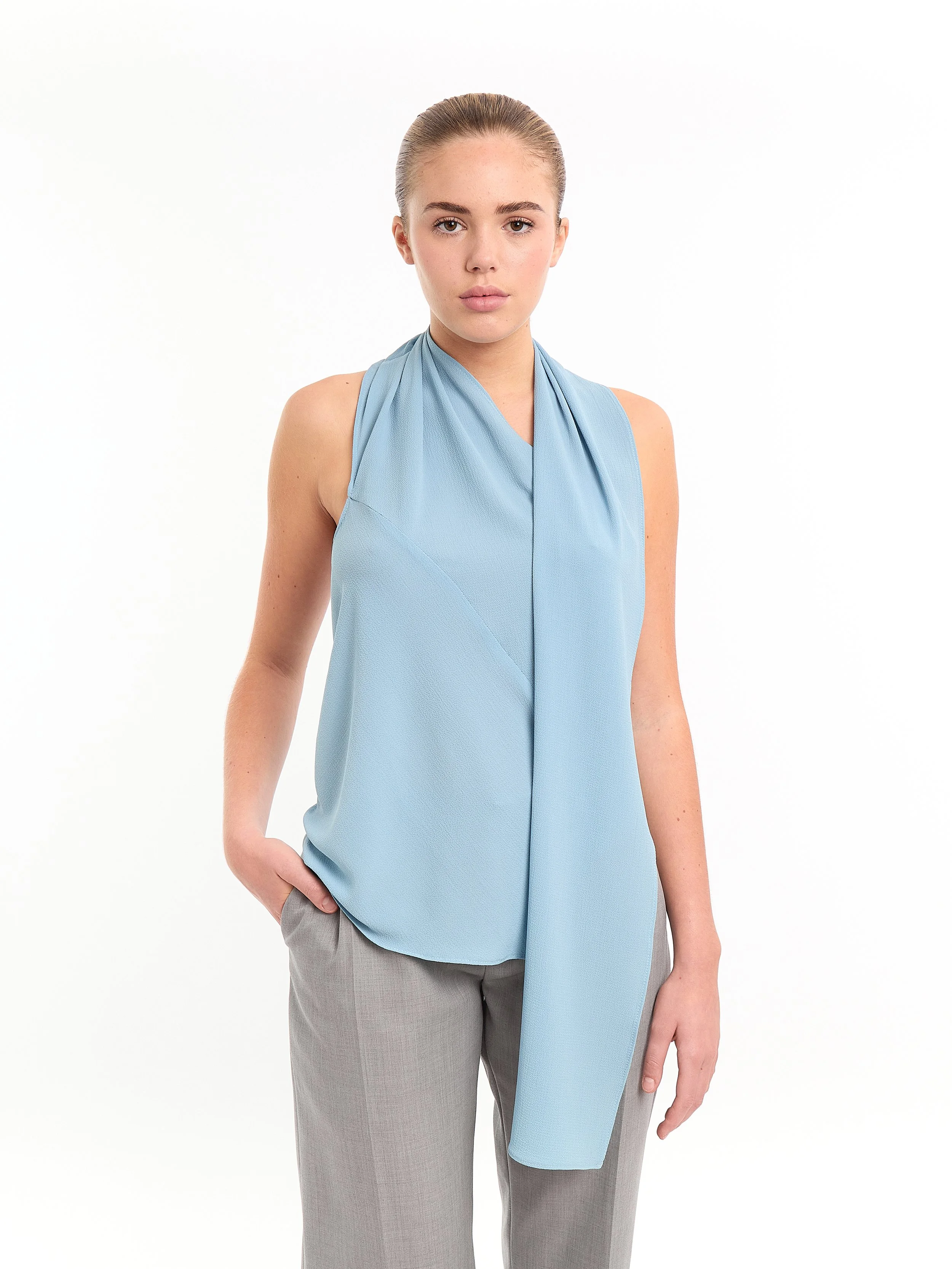 A young woman with fair skin and brown hair styled in an updo, standing with her left hand in her pocket, wearing a sleeveless, light blue blouse with a draped scarf-like detail, and light gray trousers, against a plain white background. m^