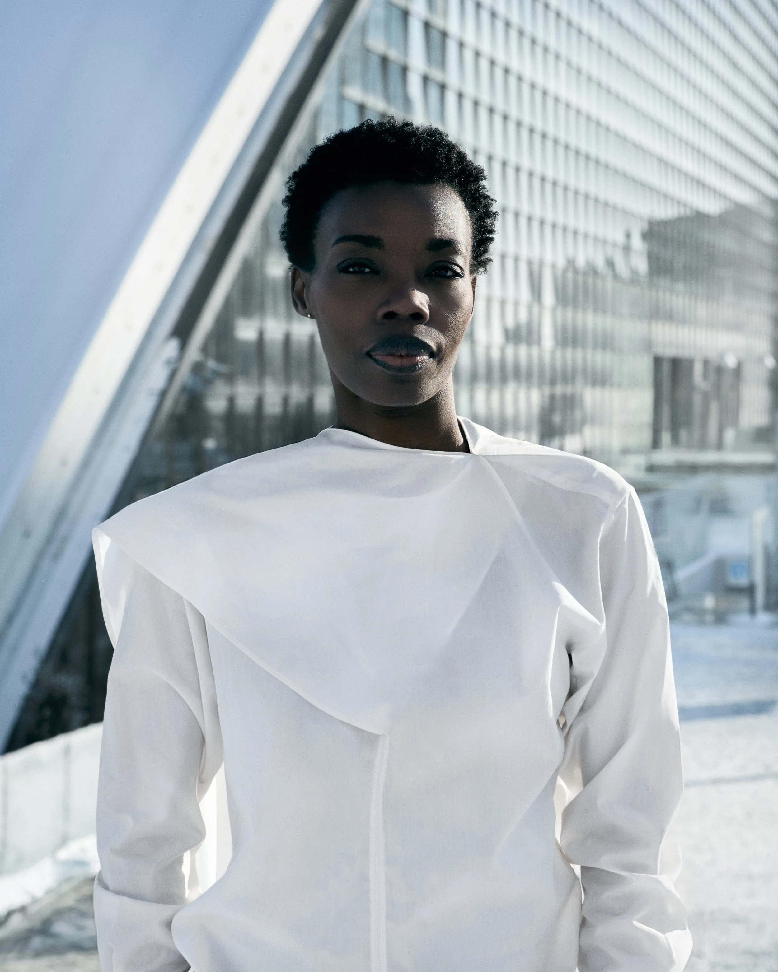 A Black woman with short curly hair wearing a white jacket, standing outside in an urban environment with modern glass buildings in the background.