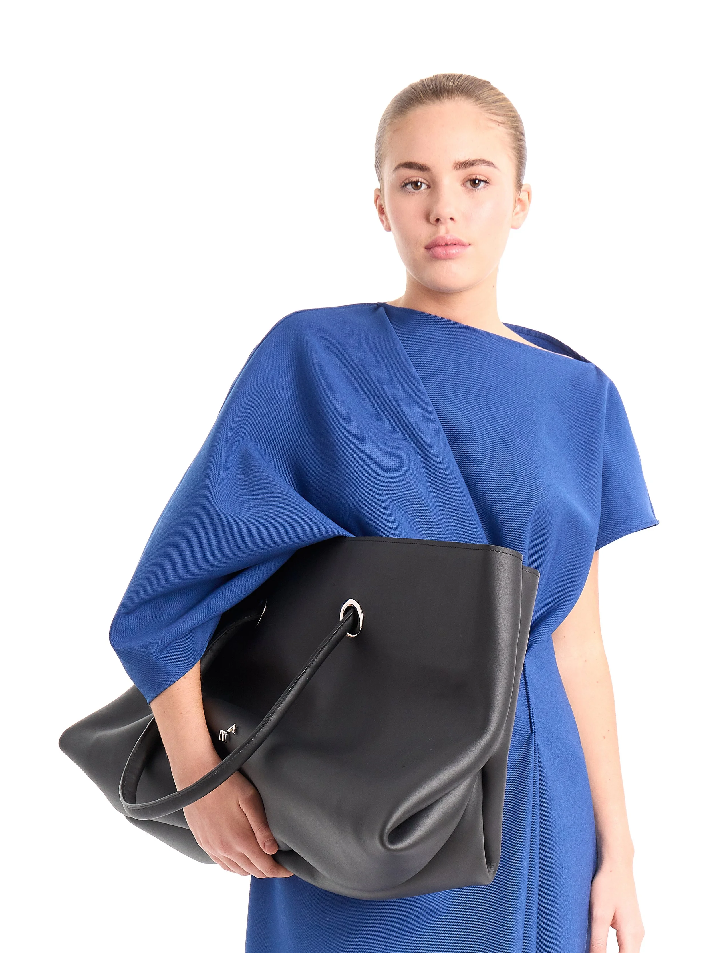 A young woman with light skin and blonde hair in a slicked-back style, wearing a blue top and carrying a large black leather handbag, standing against a plain white background. m^