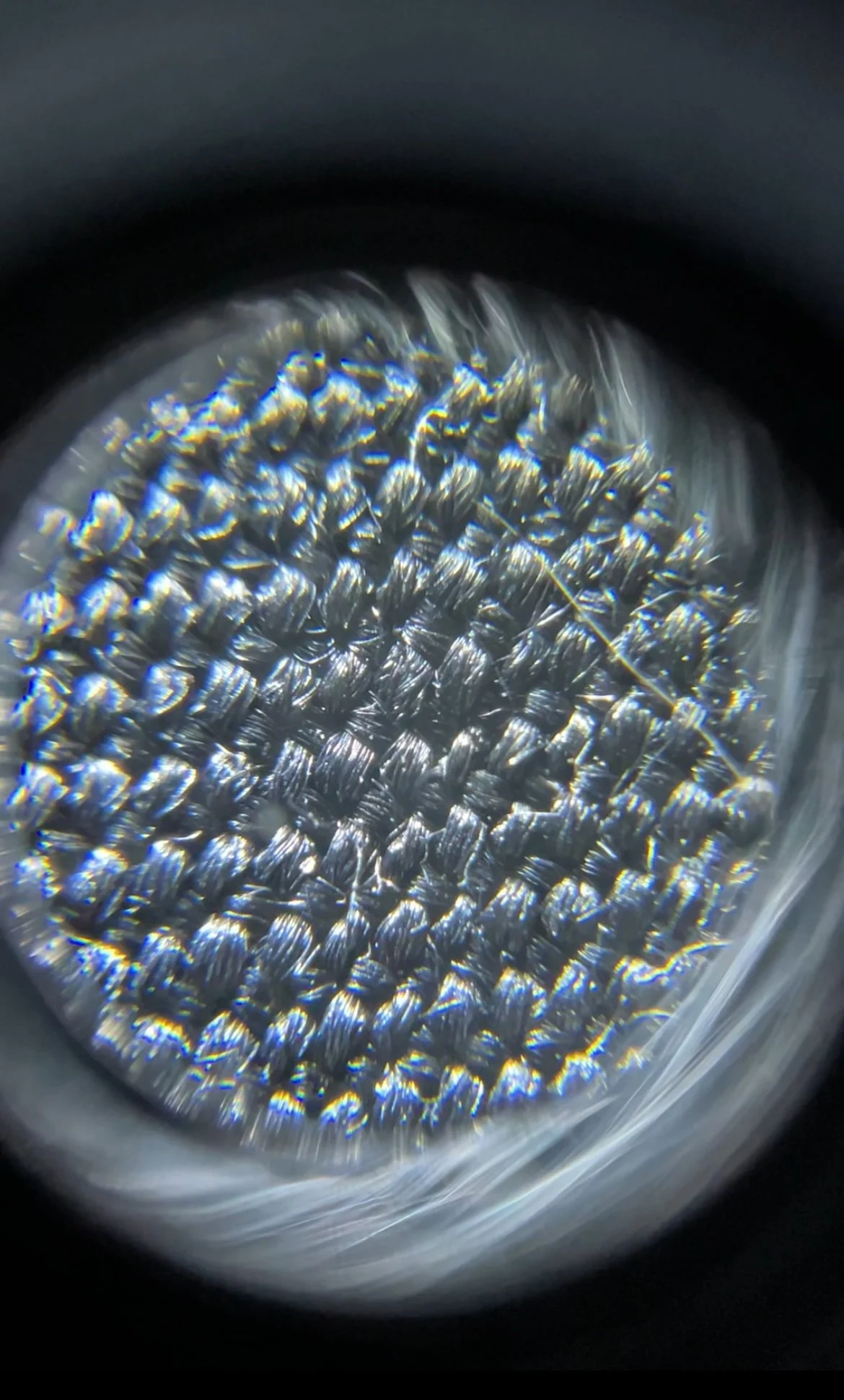 Close-up of a textured metallic surface seen through a magnifying glass, showing shiny, interwoven fibers.