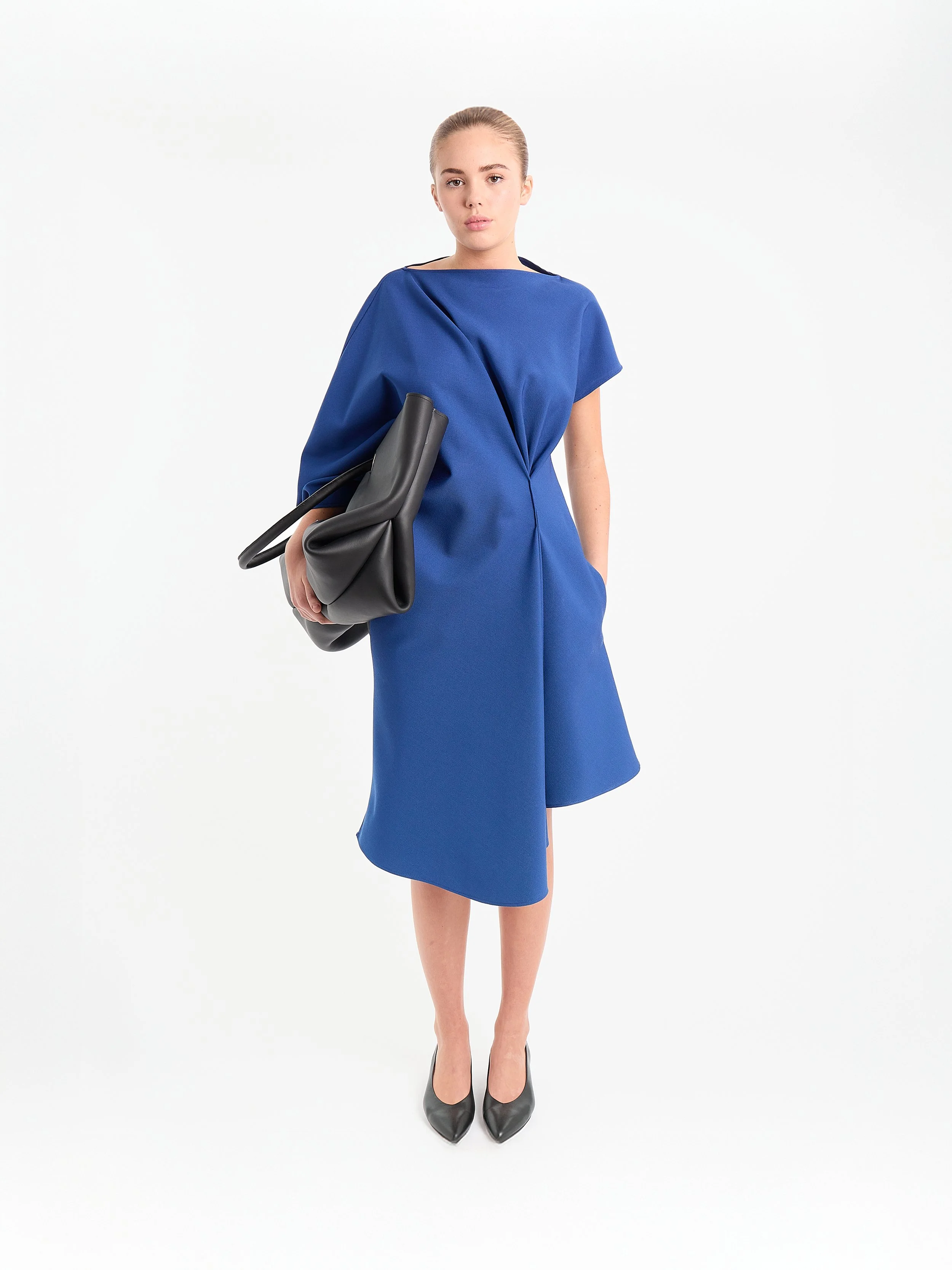 Woman wearing a blue dress, holding a black leather bag, standing against a plain white background. A fluid, relaxed silhouette cut in advanced fabric — waterproof and wrinkle-resistant. Engineered for freedom, it drapes with ease while hidden pocket