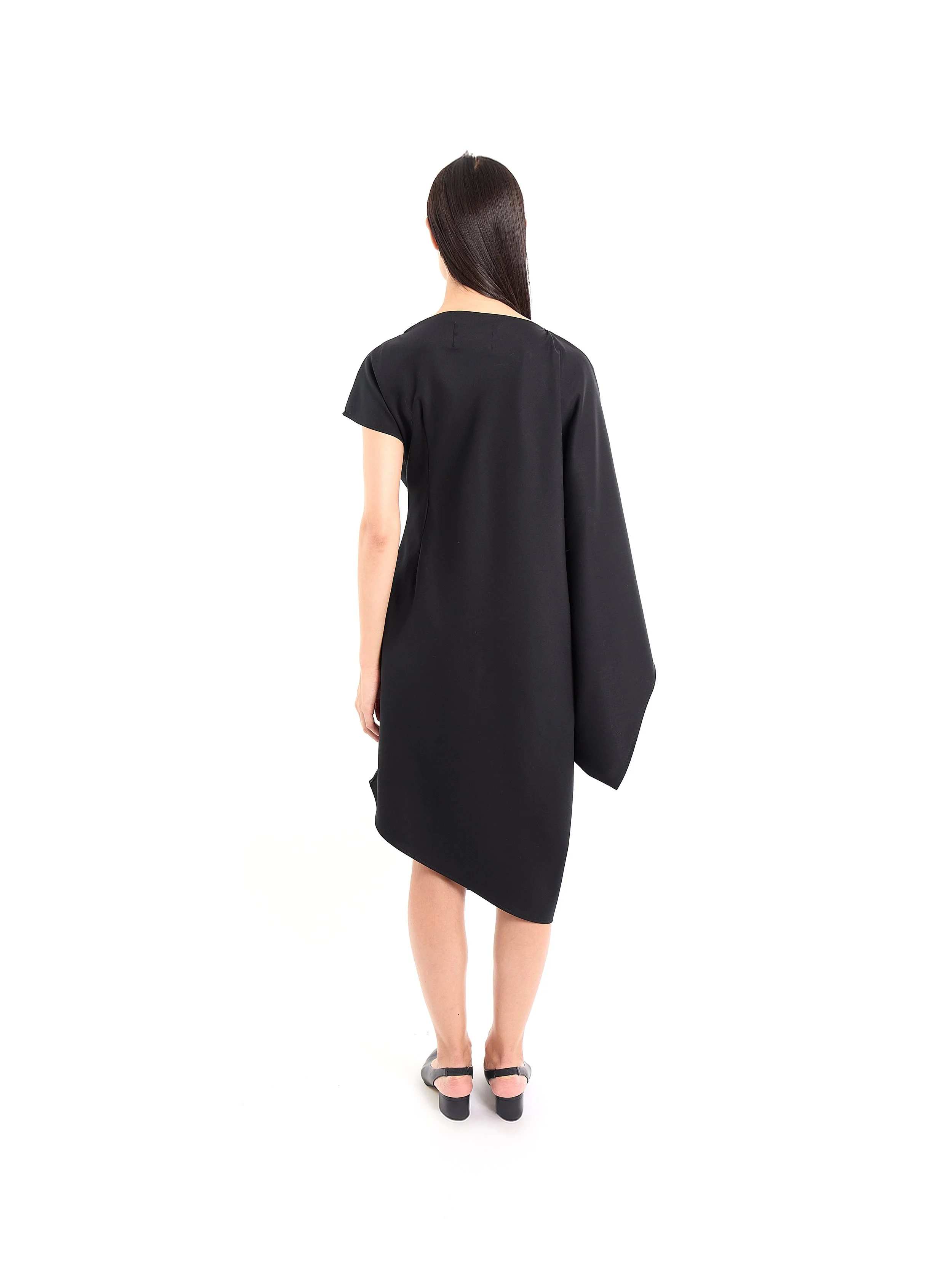 Back view of a woman with long dark hair wearing a black knee-length dress, holding a matching black jacket, standing barefoot on a white background. m^