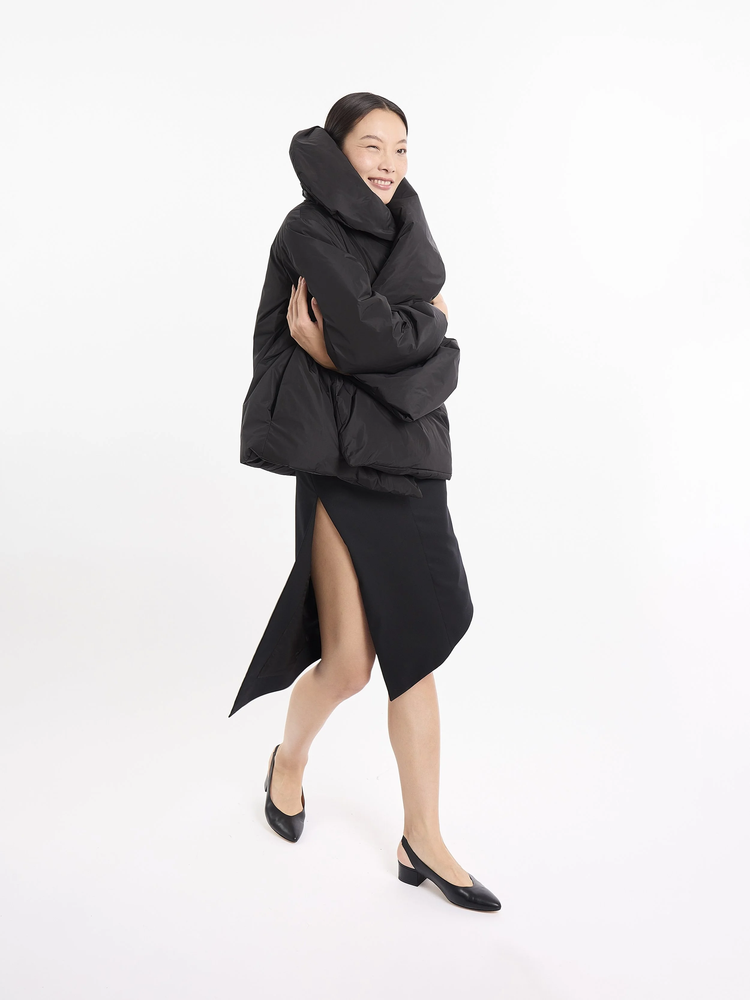 A woman posing in a mpower black puffer jacket, black high-low skirt with a slit, and black heels, standing against a white background.