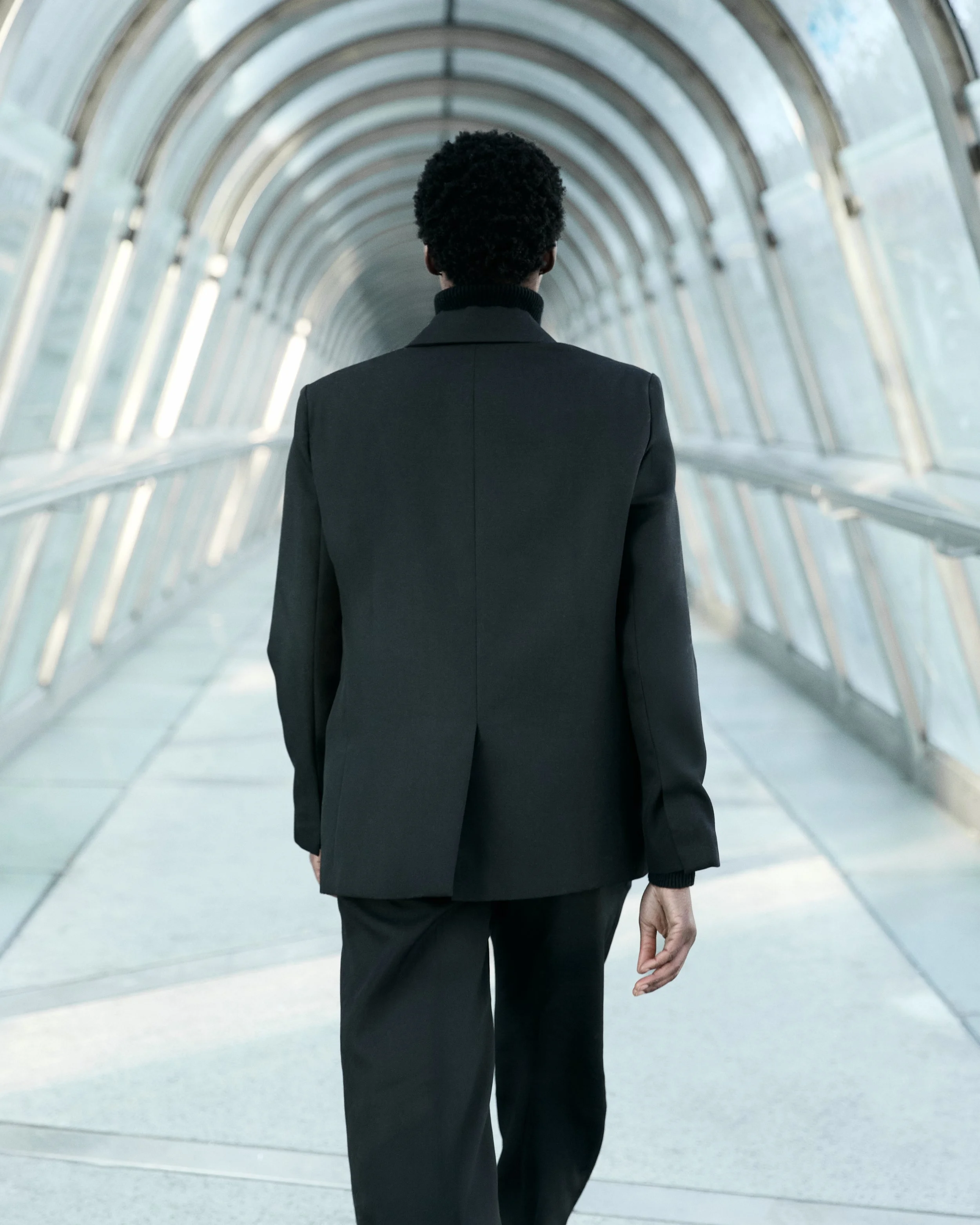 A person in a black suit walking through a glass enclosed pedestrian bridge.