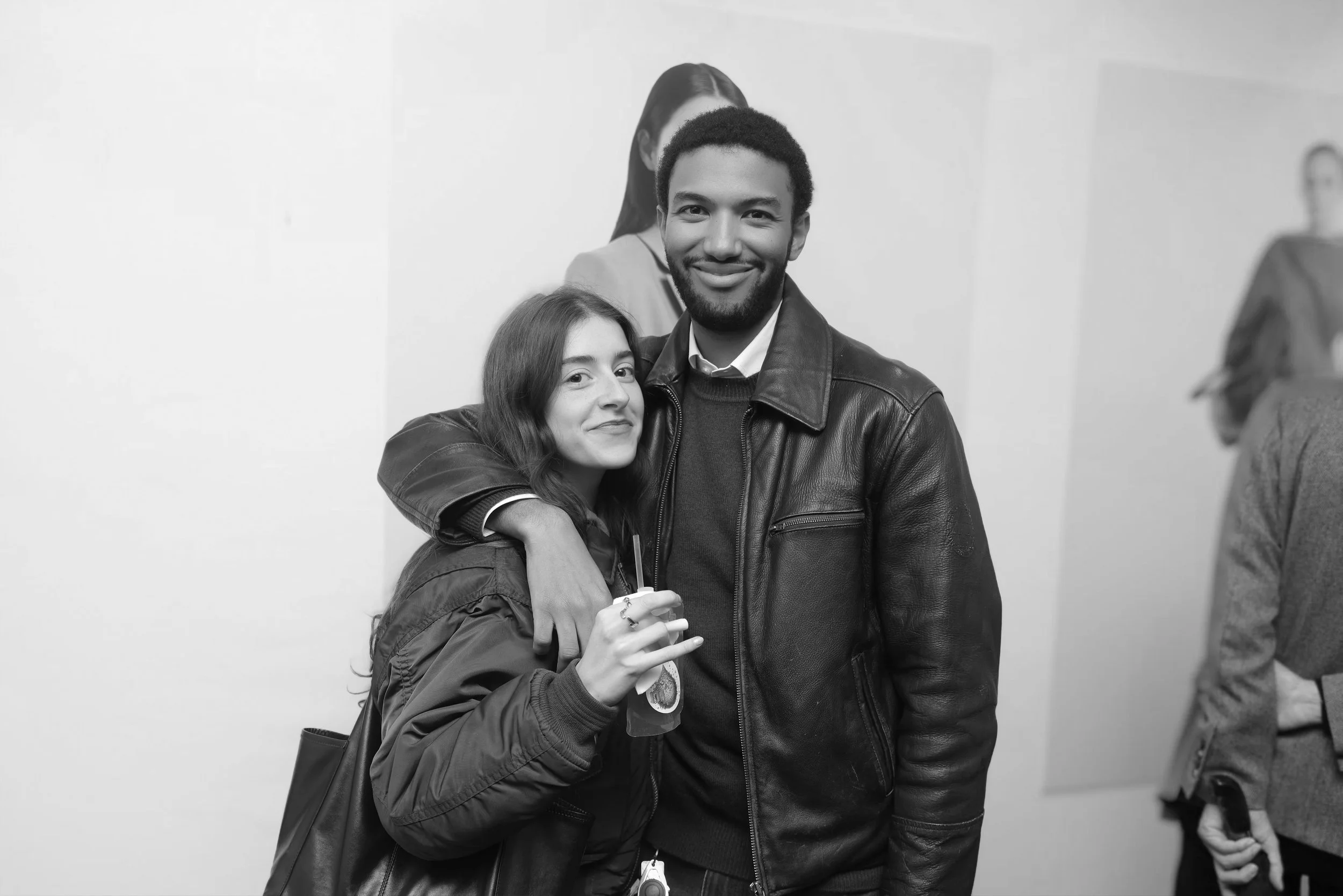 Two young adults, a woman and a man, smiling and posing together at an indoor event, with other people in the background.