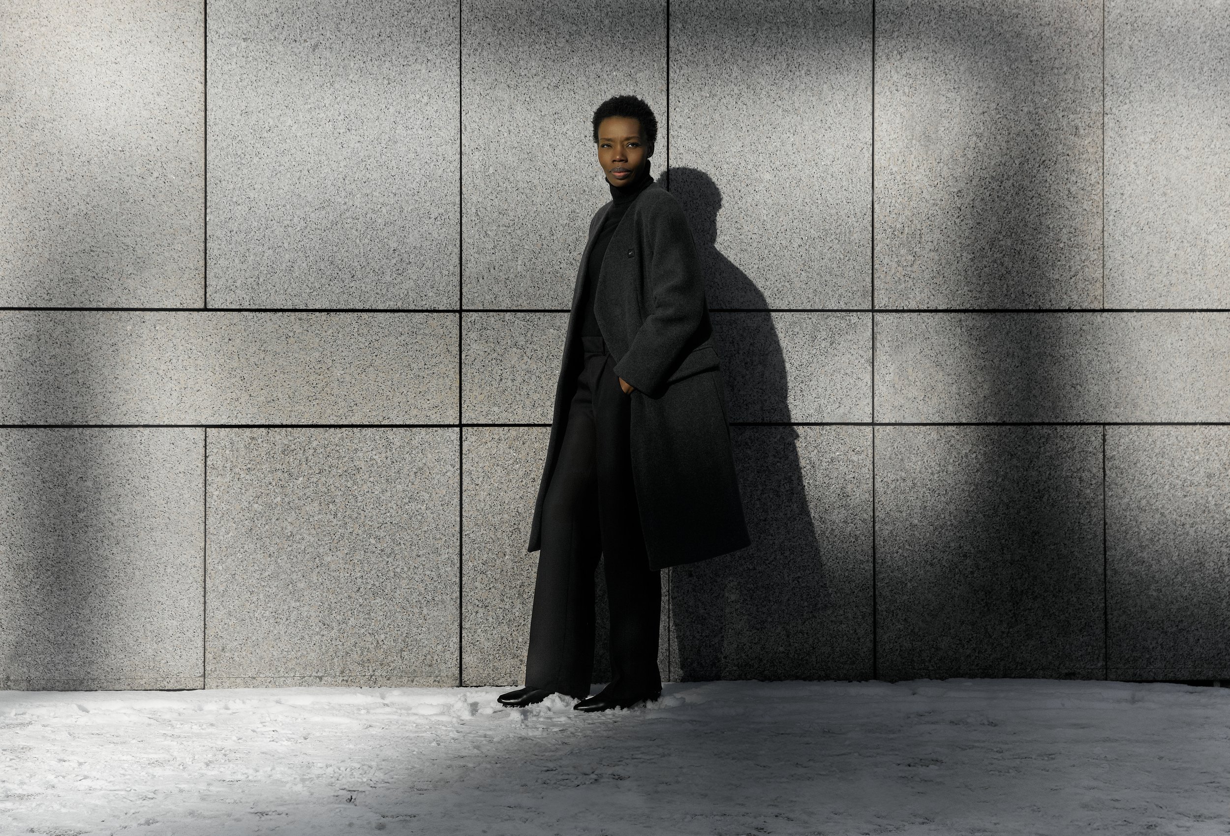 A woman with short curly hair wearing a long dark coat, black pants, and black shoes standing against a large grey tiled wall, casting a shadow on the wall in bright sunlight.