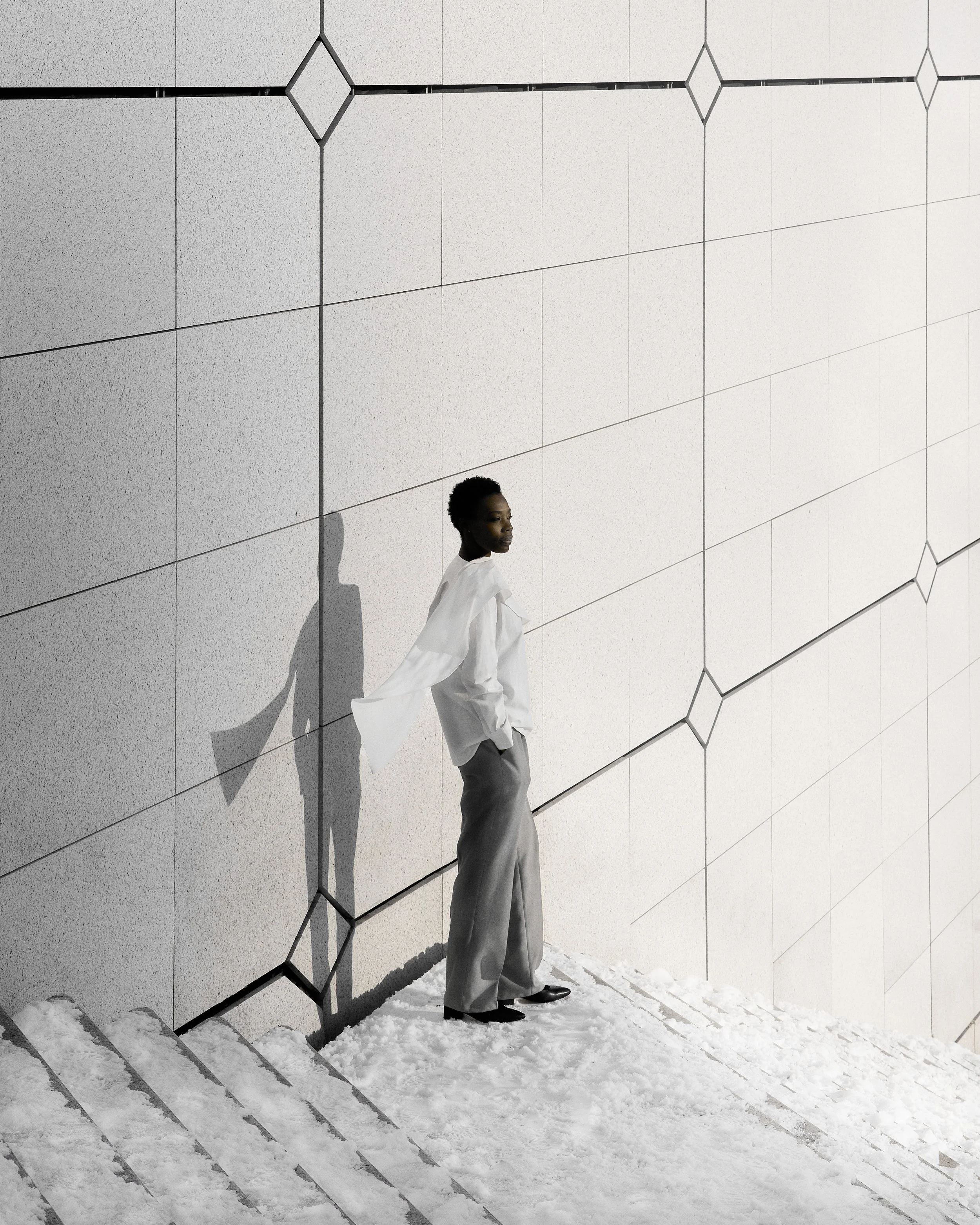 A woman standing on snow-covered ground against a geometrically patterned wall with her shadow cast on the wall.