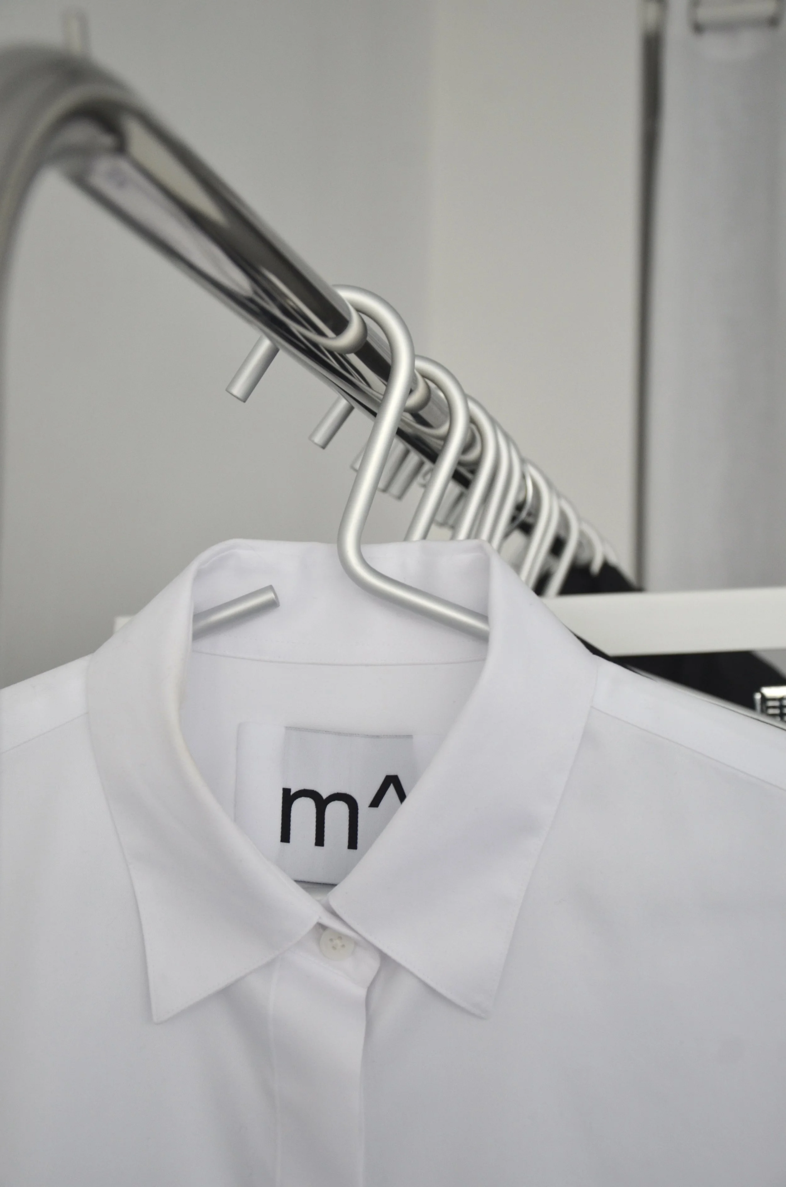 White dress shirt hanging on a metal clothing rack with several other hangers visible in the background. m^ready to wear showroom.