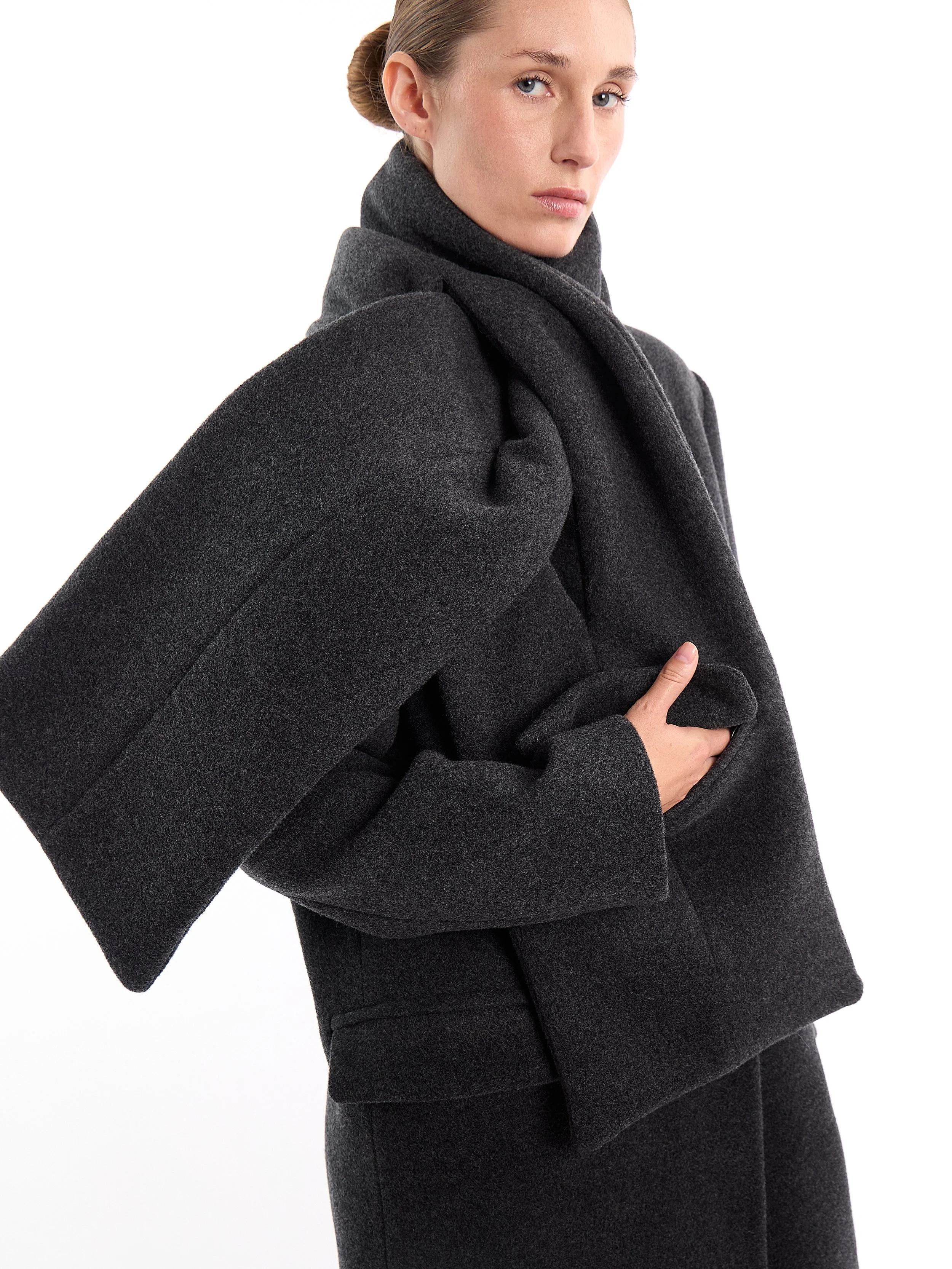 A woman in a dark gray coat with a large collar and matching pants, standing against a white background. m^