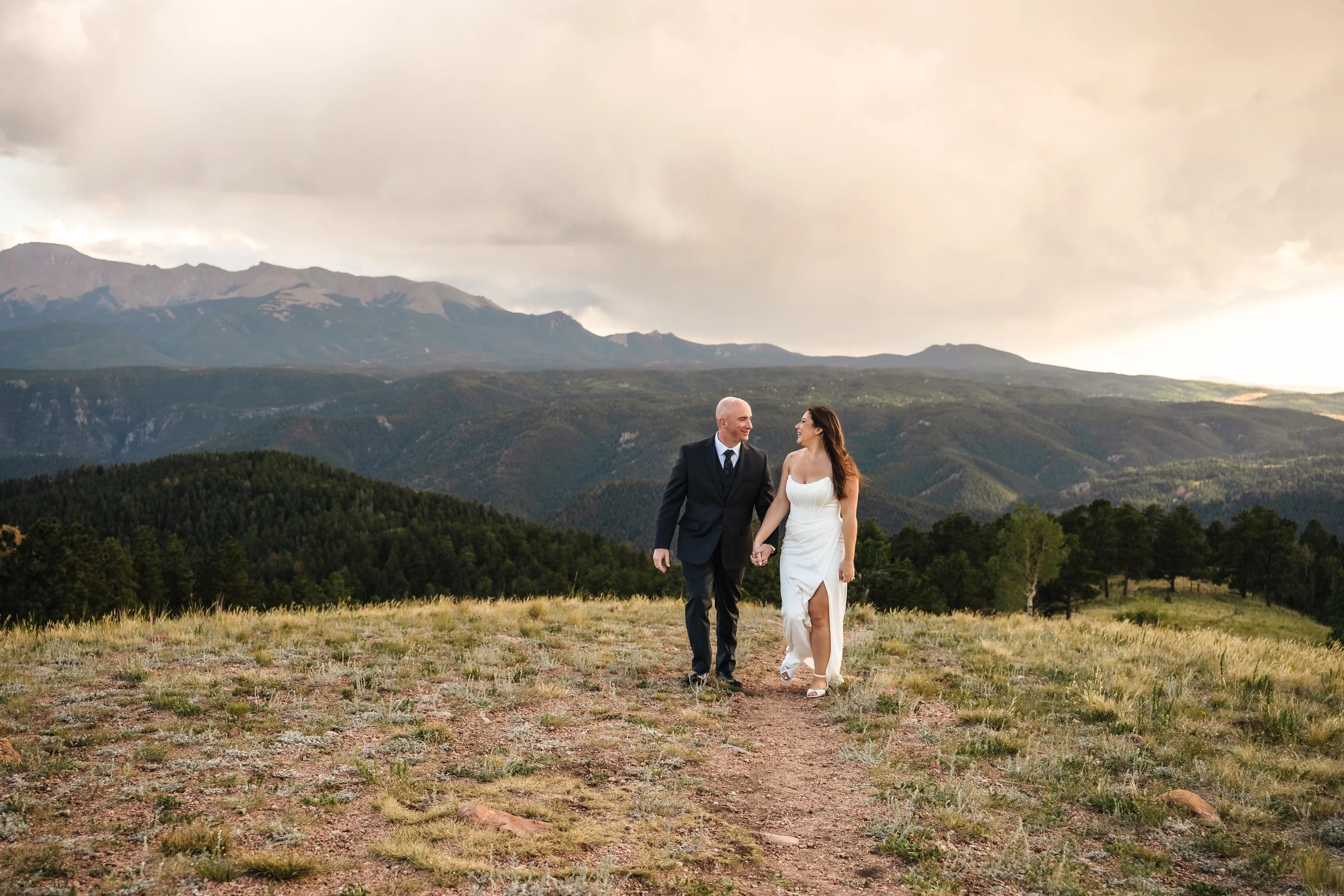 Bride and groom walking hand in hand during an intimate Colorado Springs elopement with panoramic views of Pikes Peak and the Front Range beneath dramatic summer storm clouds. Captured by Colorado Springs elopement photographer Sarah K. Dunn.