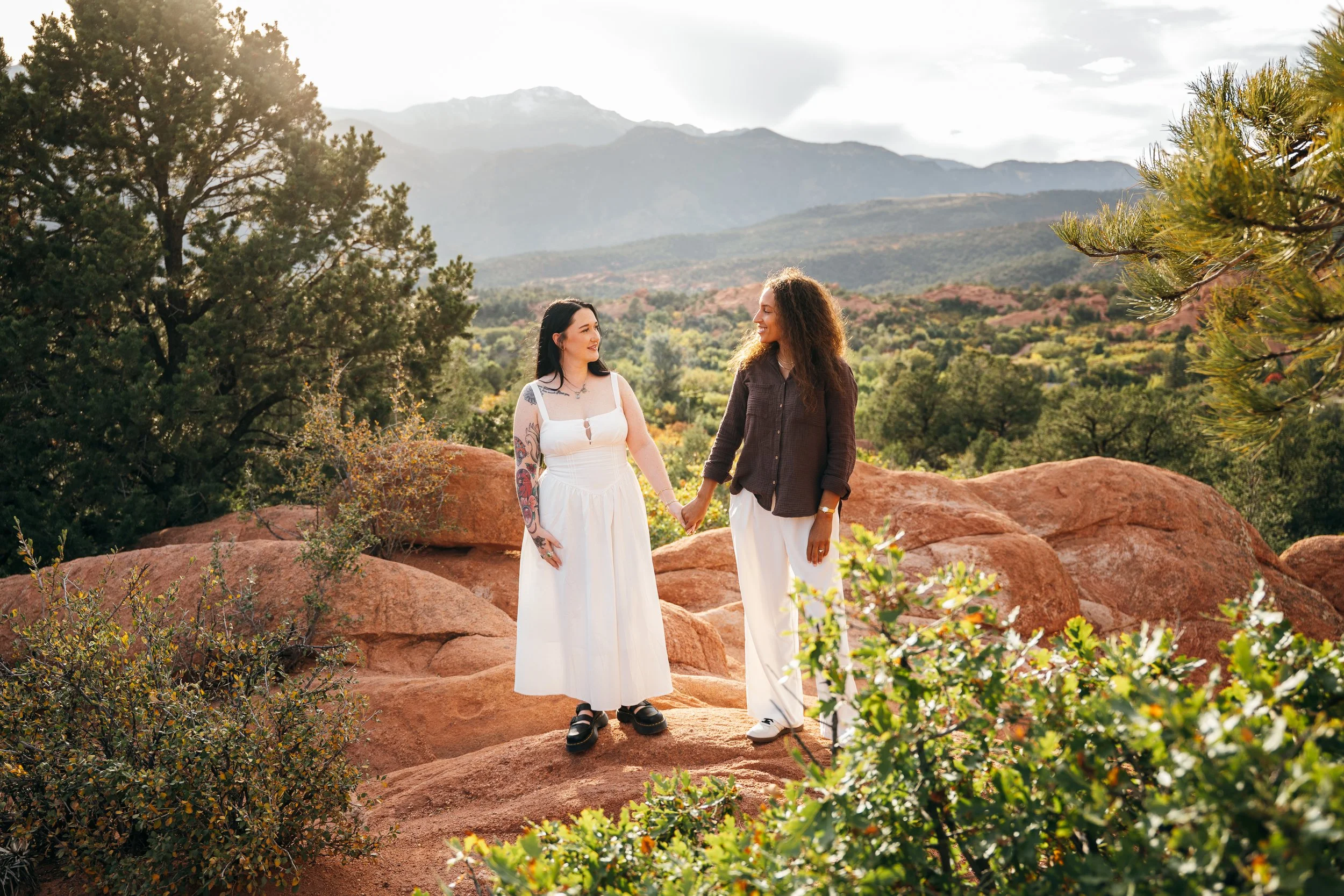Two women holding hands on red rocks with Pikes Peak and green foothills behind them.