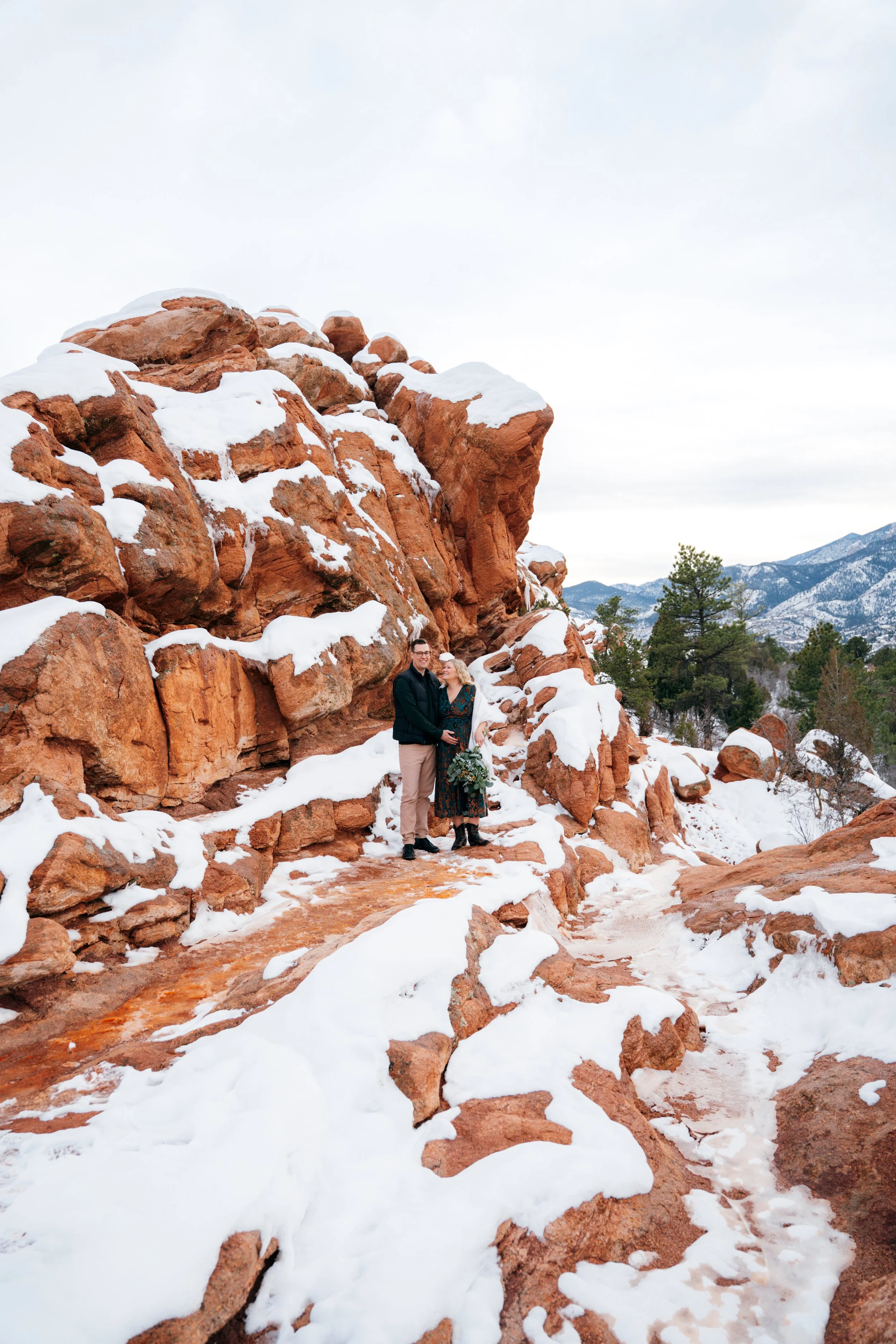 Couple standing among snow-covered red rocks during a winter maternity session at Garden of the Gods in Colorado Springs.