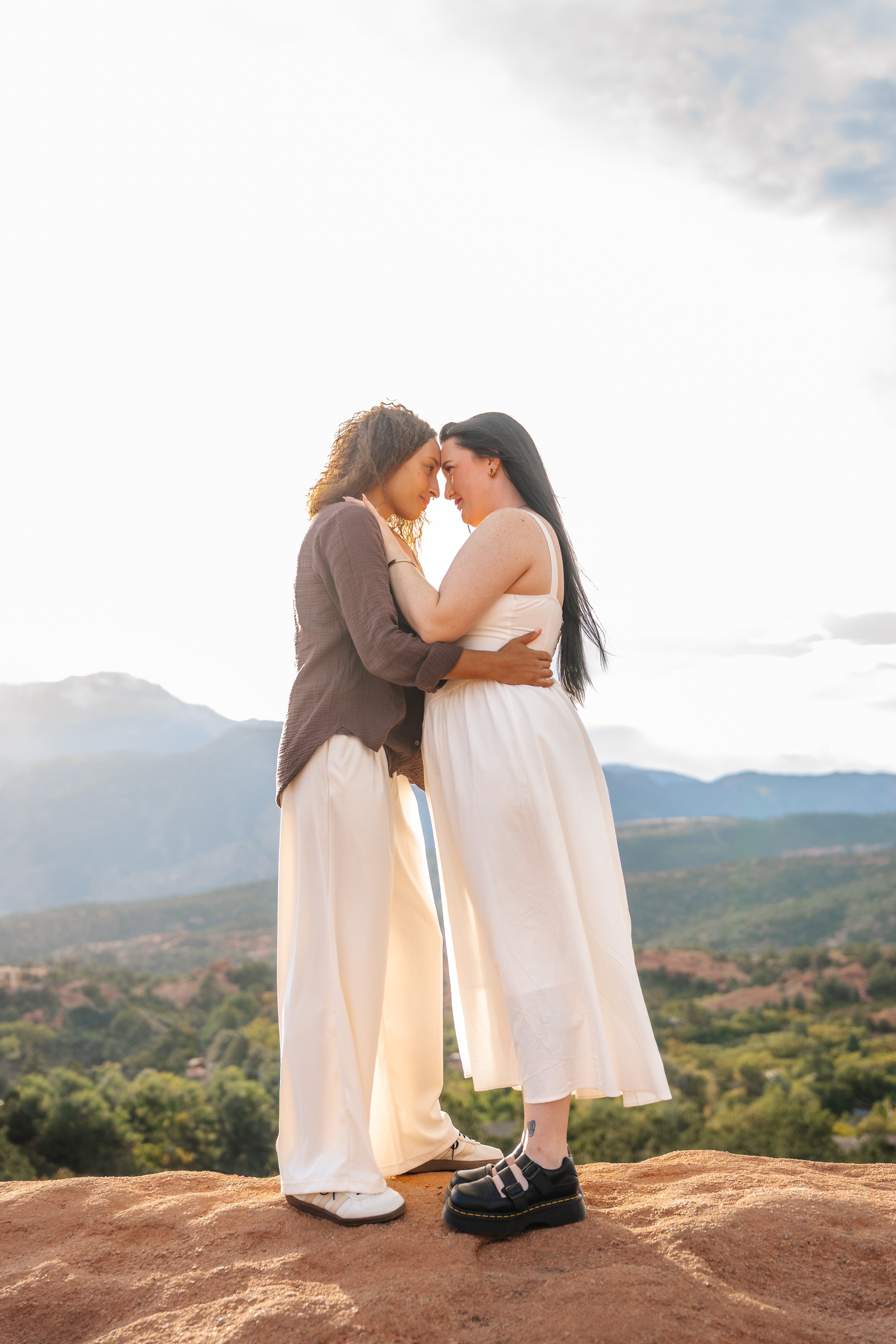 Two women standing close with their foreheads touching on red sandstone overlooking the mountains.
