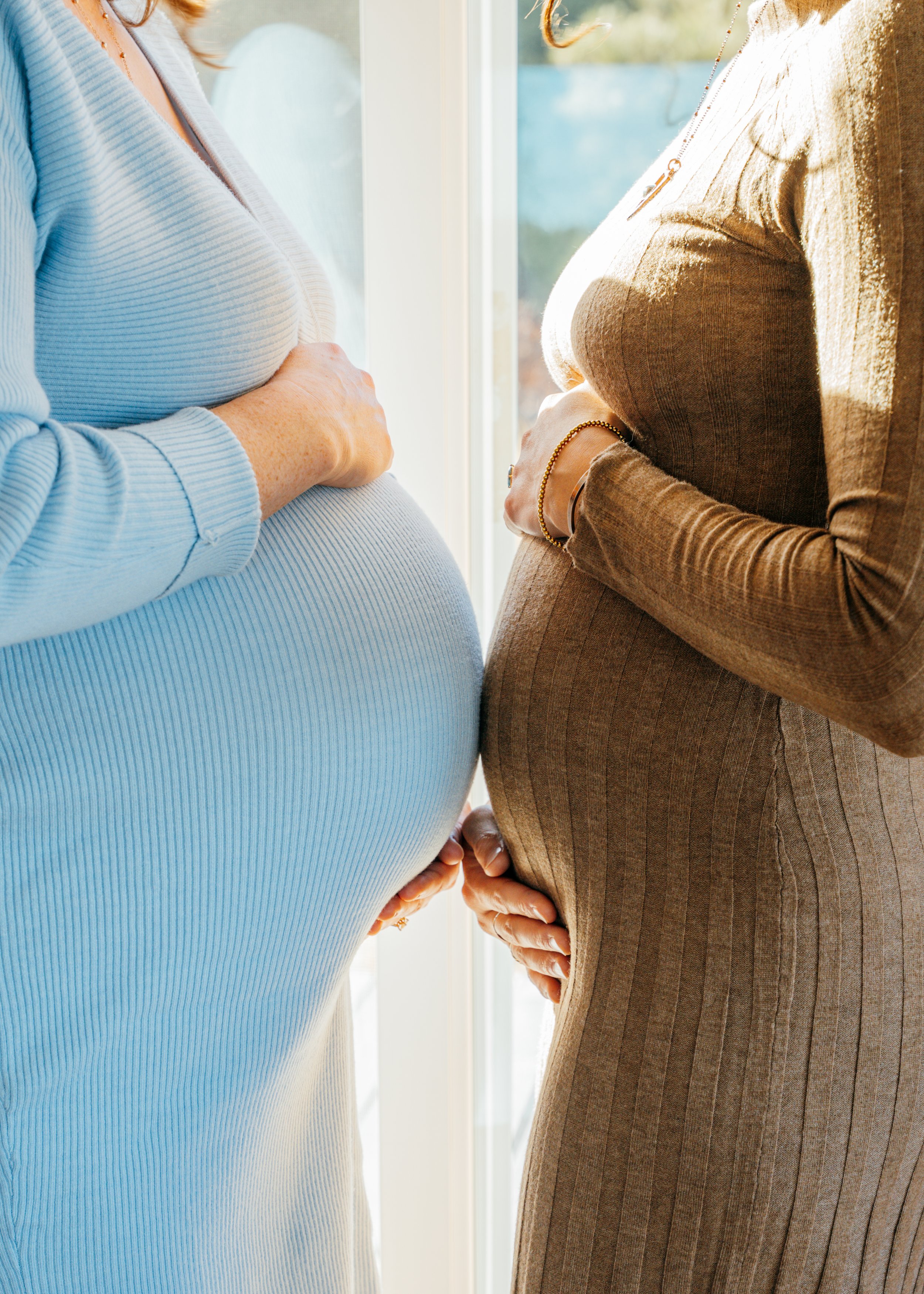 Two pregnant women standing together by a window during a cozy in-home maternity session in Colorado Springs