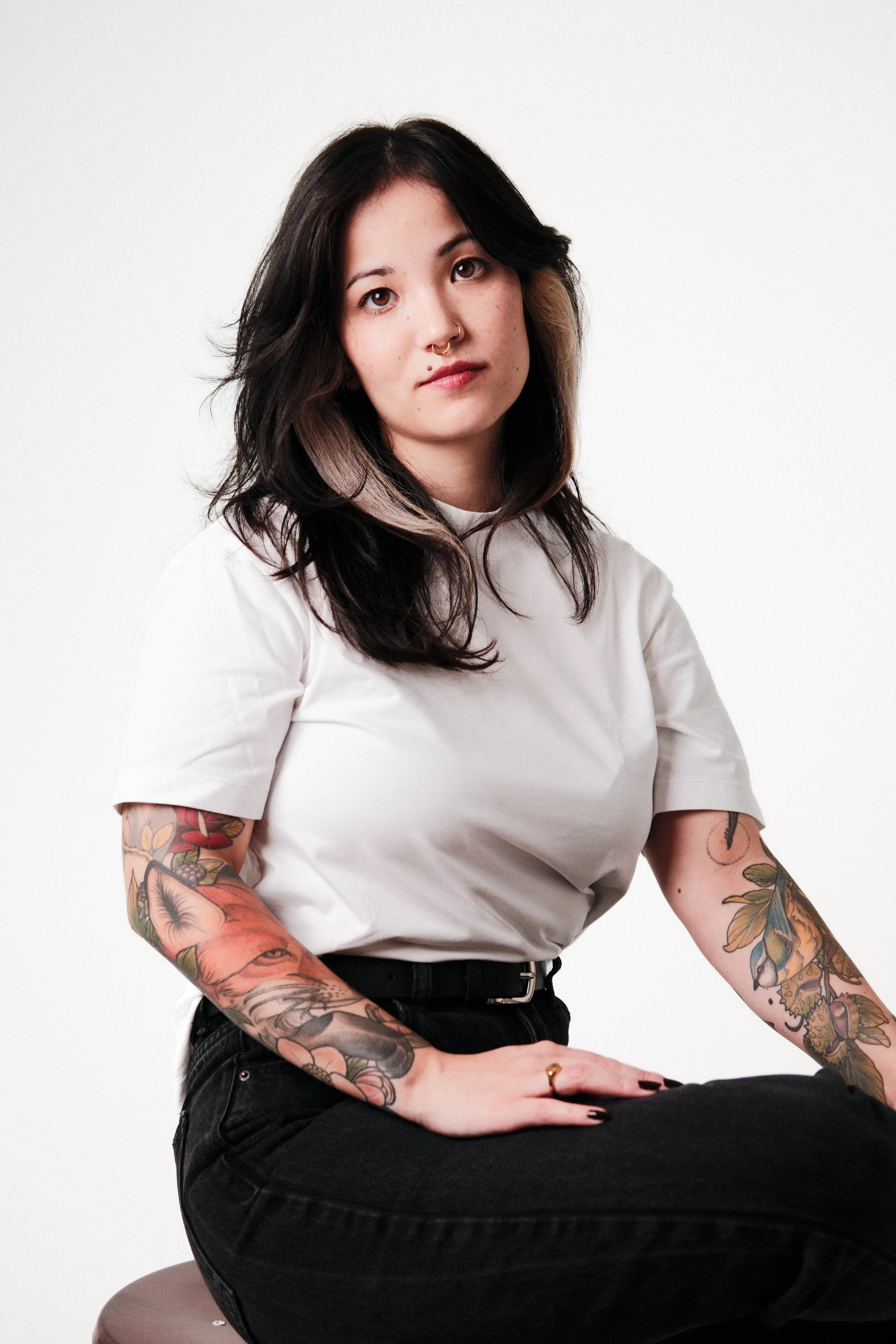 Junge Frau mit Tattoos, schwarze Haare mit einem hellen Fleck, in weißem T-Shirt sitzend vor weißem Hintergrund.