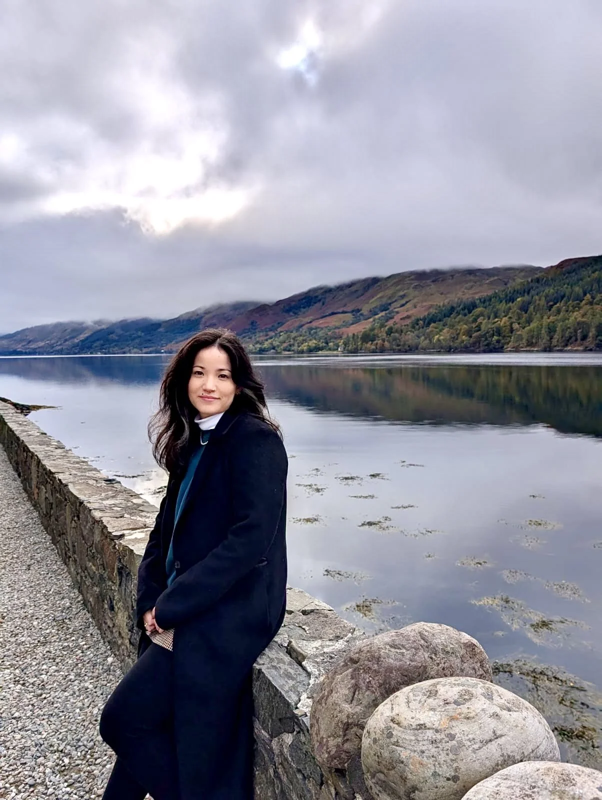 Eine junge Frau steht an einer Steinmauer am Ufer eines Sees mit Bergen im Hintergrund, bewölkter Himmel.
