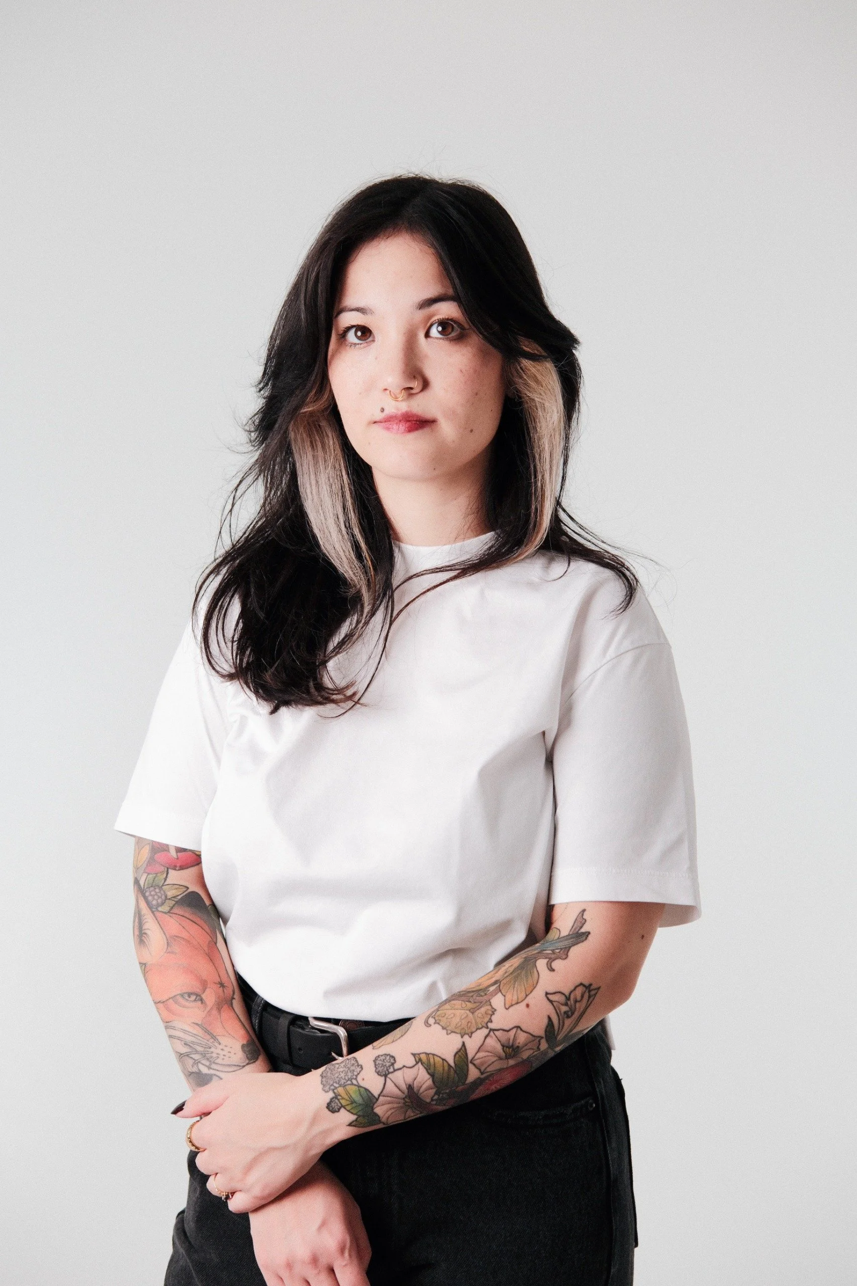 Junge Frau mit schwarzen und blonden Haaren, trägt ein weißes T-Shirt und schwarze Hose, hat tätowierte Arme, steht vor hellem Hintergrund.