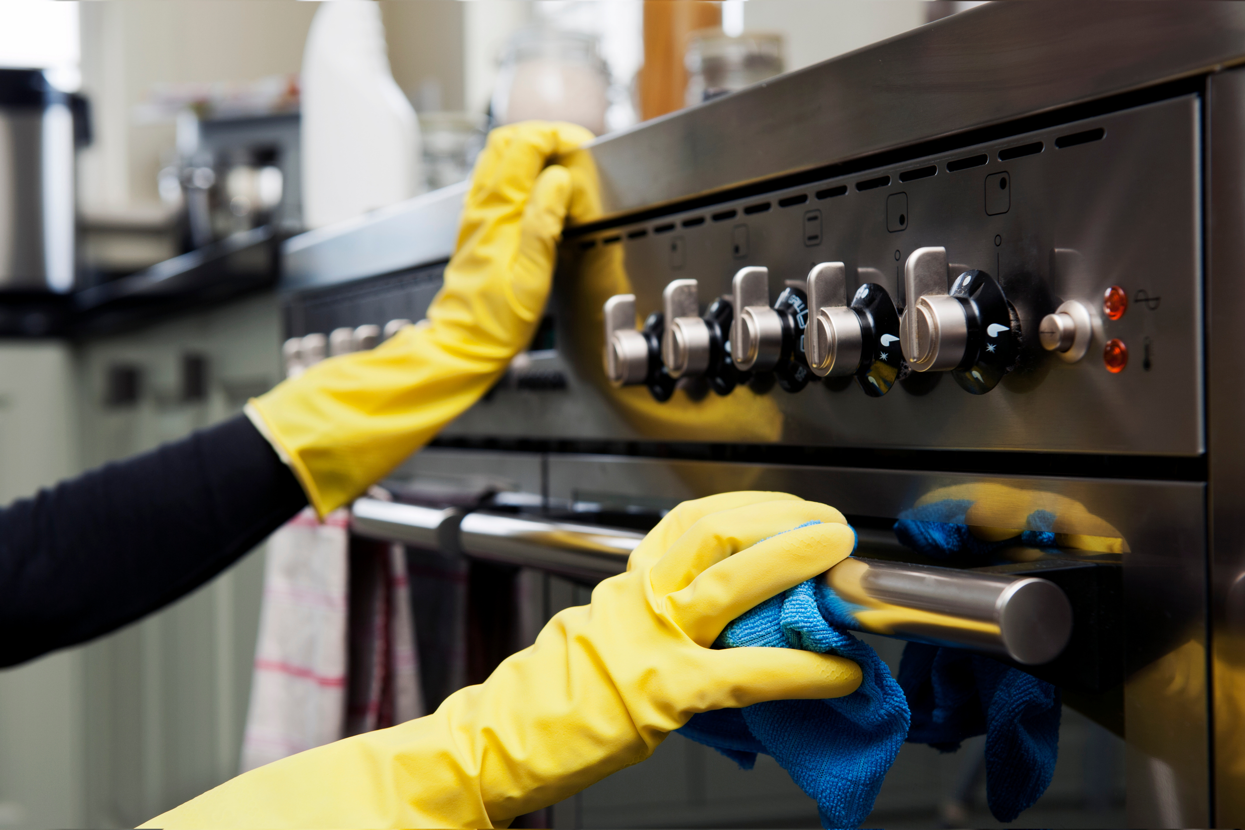 Person wearing yellow rubber gloves cleaning a stainless steel oven with a blue cloth in a kitchen.