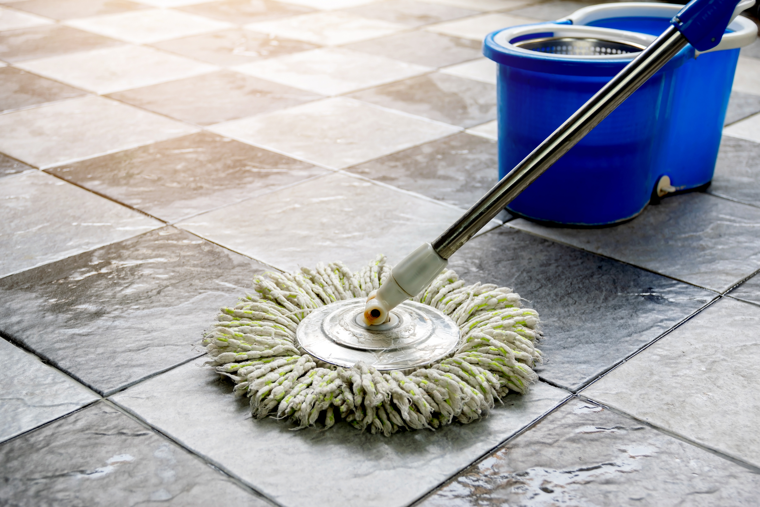 A mop cleaning a tiled floor with a blue bucket nearby.