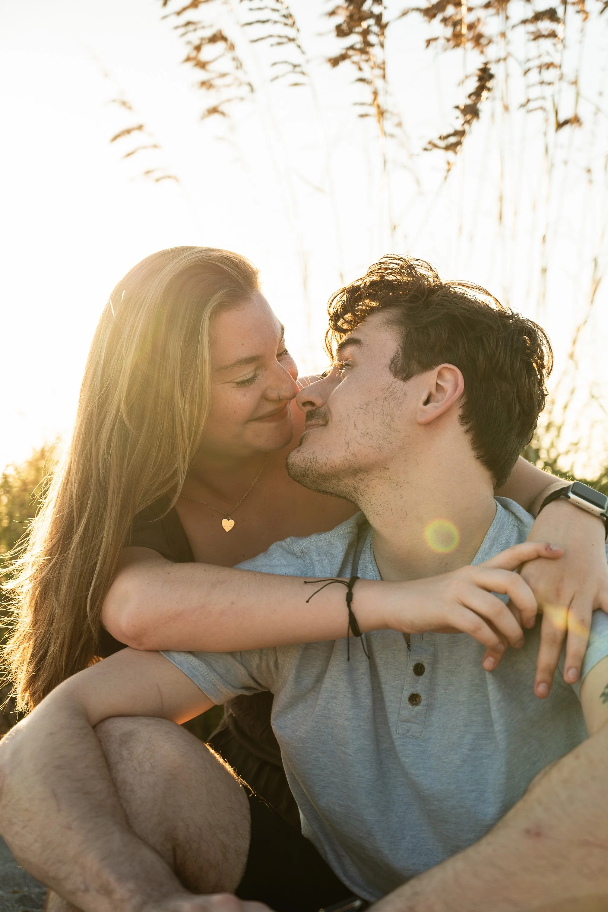 A young couple is outdoors during sunset, embracing and smiling as they look at each other closely, with trees in the background and warm sunlight highlighting their faces.