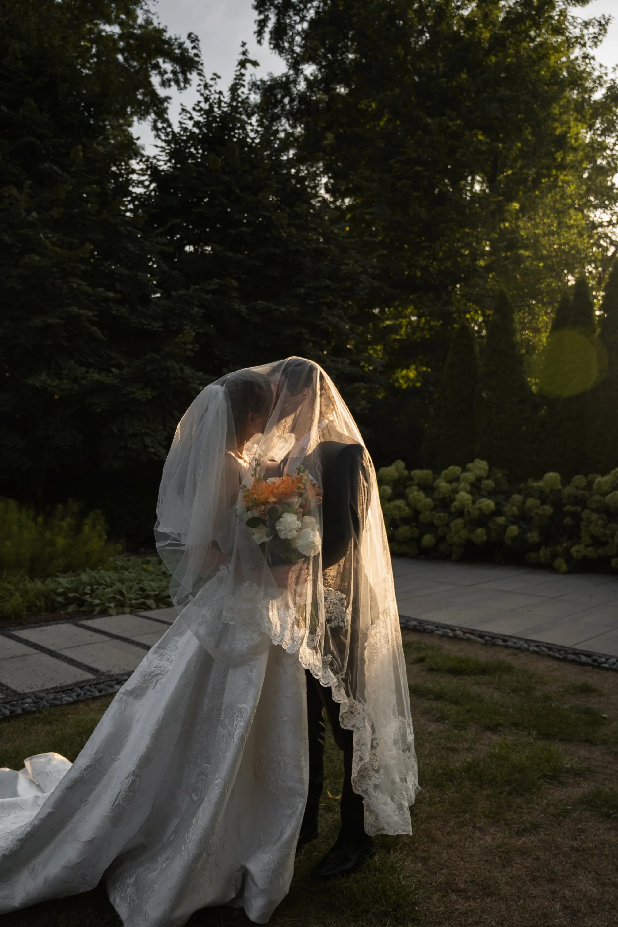 A bride dressed in a white wedding gown and veil holding a bouquet of flowers, standing outdoors with trees and flowers in the background, illuminated by sunlight.