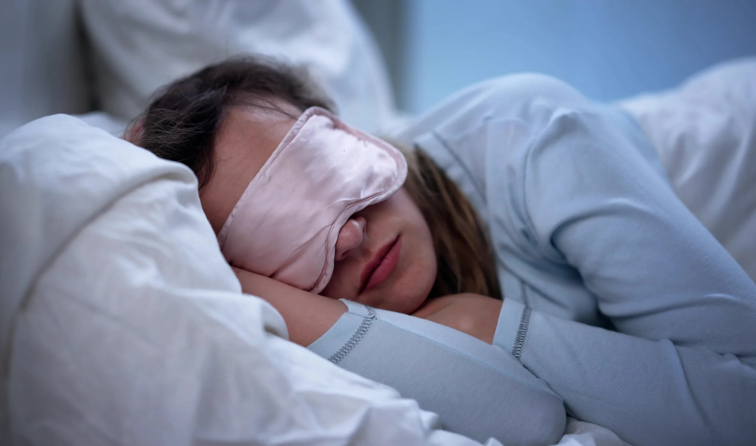 A woman sleeping peacefully with a pink satin sleep mask covering her eyes, lying on a bed with white sheets and pillowcases, wearing a light-colored long-sleeve shirt.