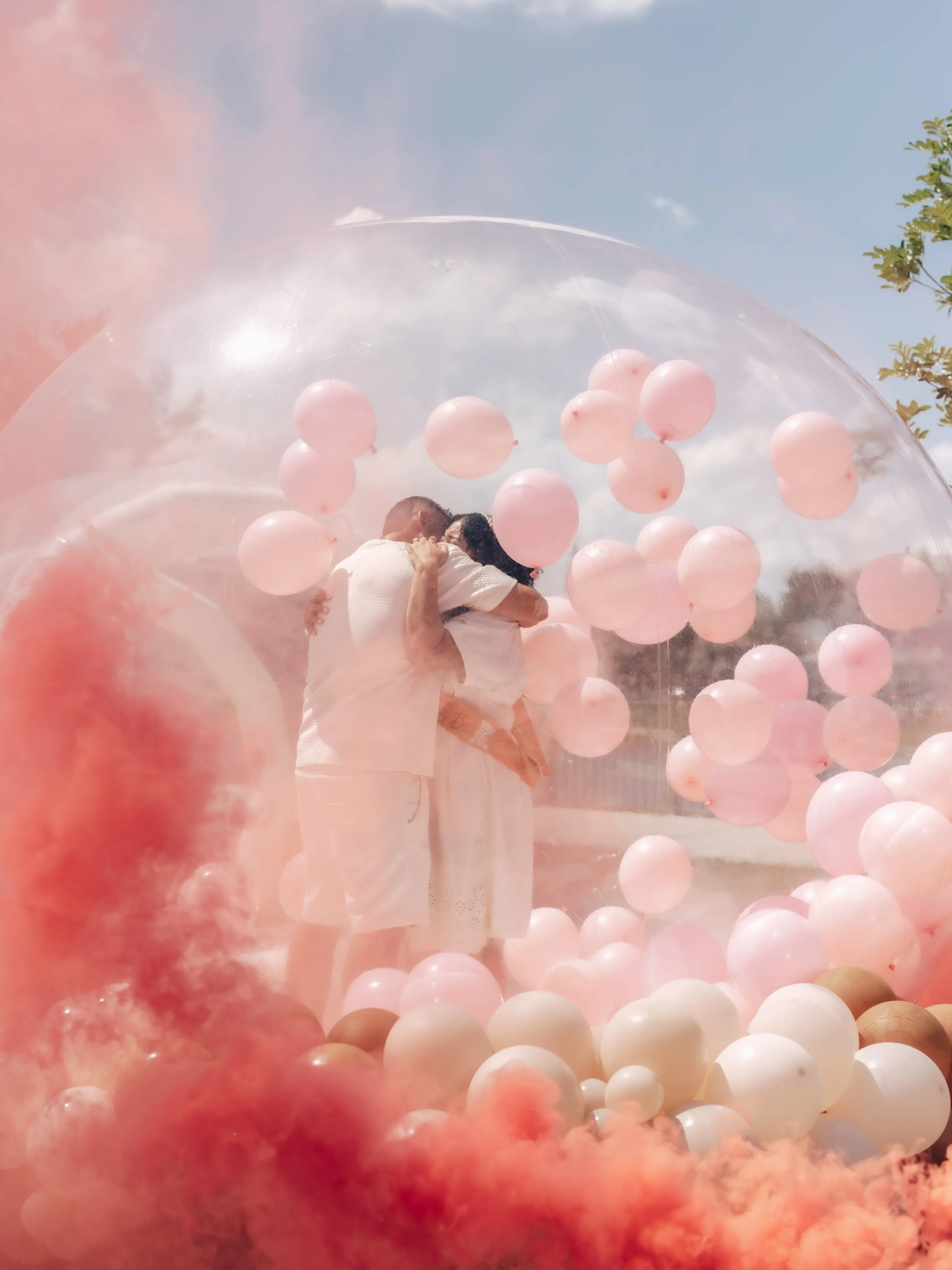 A couple embraces inside a transparent balloon filled with pink balloons, with a cloudy sky and green tree in the background.