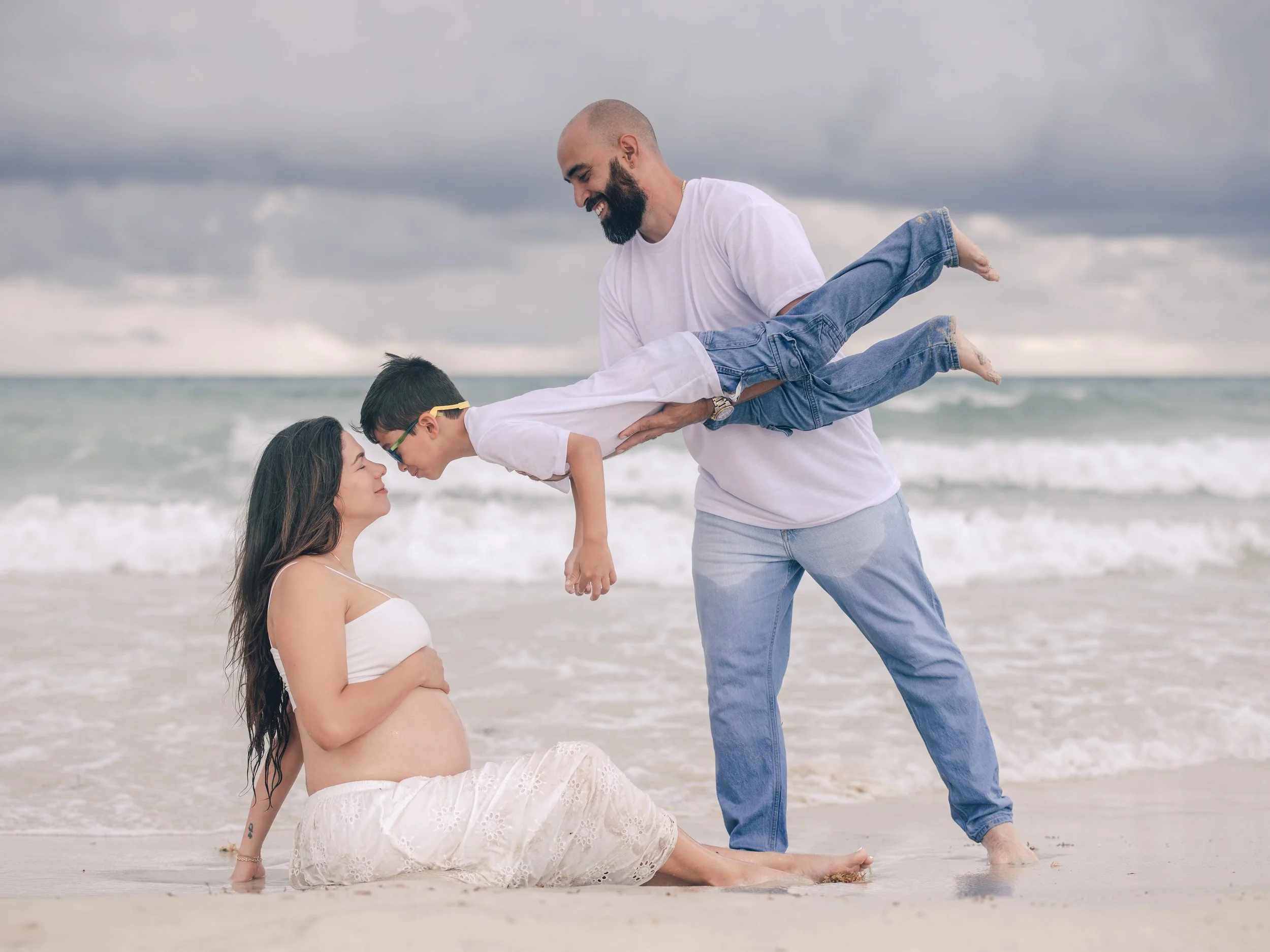 A family of three, with one woman sitting on the sand and two males, possibly a father and son, standing in the ocean waves at the beach. They are celebrating, with the woman smiling and the males in a playful pose.