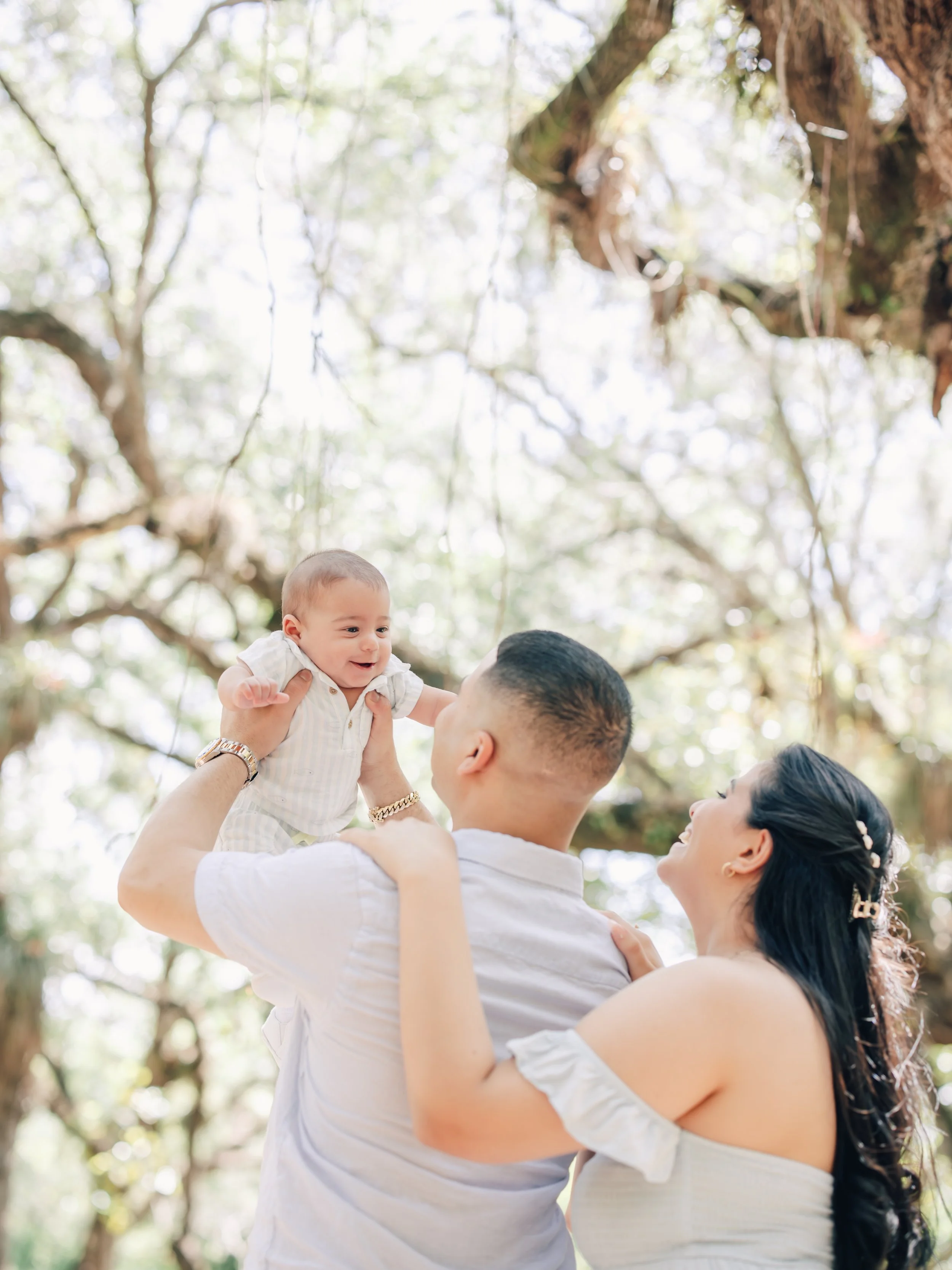 A happy family outdoors with a father lifting a baby in the air, surrounded by trees and sunlight.