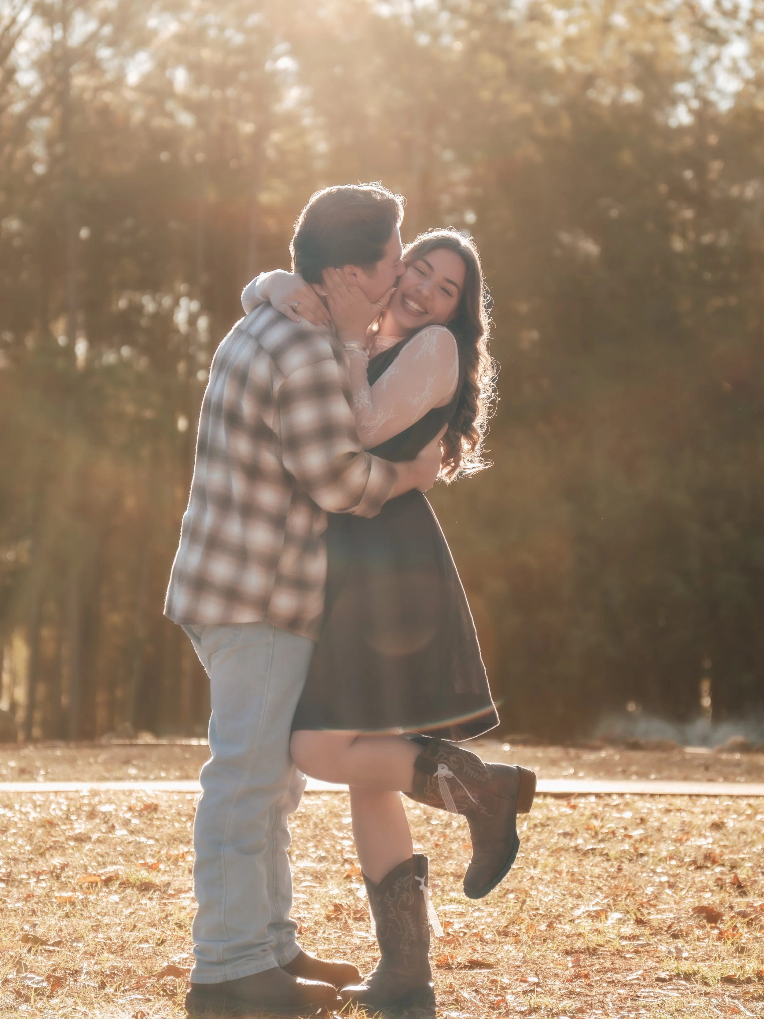 A happy couple is outdoors in a park in central florida during sunset, embracing and kissing each other. The woman is standing on one leg, with the other lifted, wearing a black dress and cowboy boots. The man is wearing a plaid shirt and jeans.