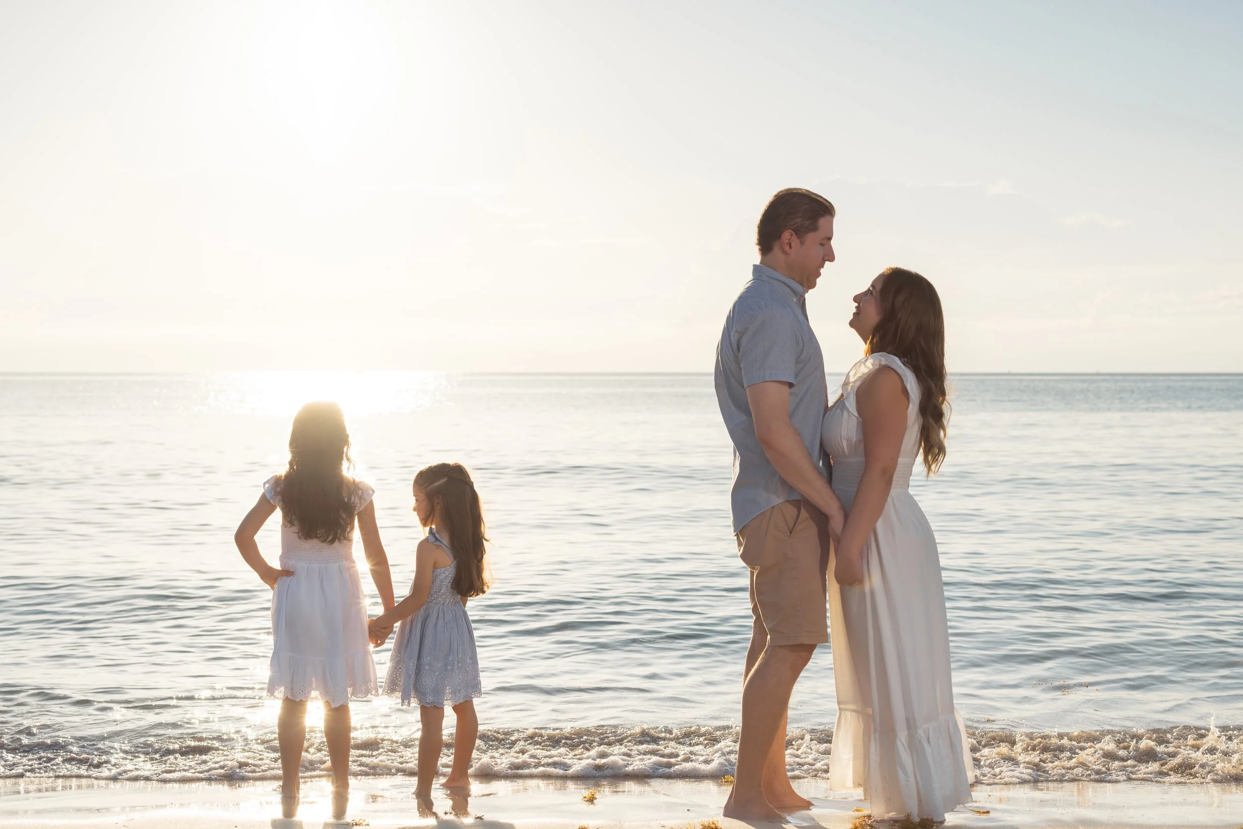 A couple stands holding hands facing each other on the beach at sunset, with three young girls nearby, some playing in the water and one with her hands on her hips.