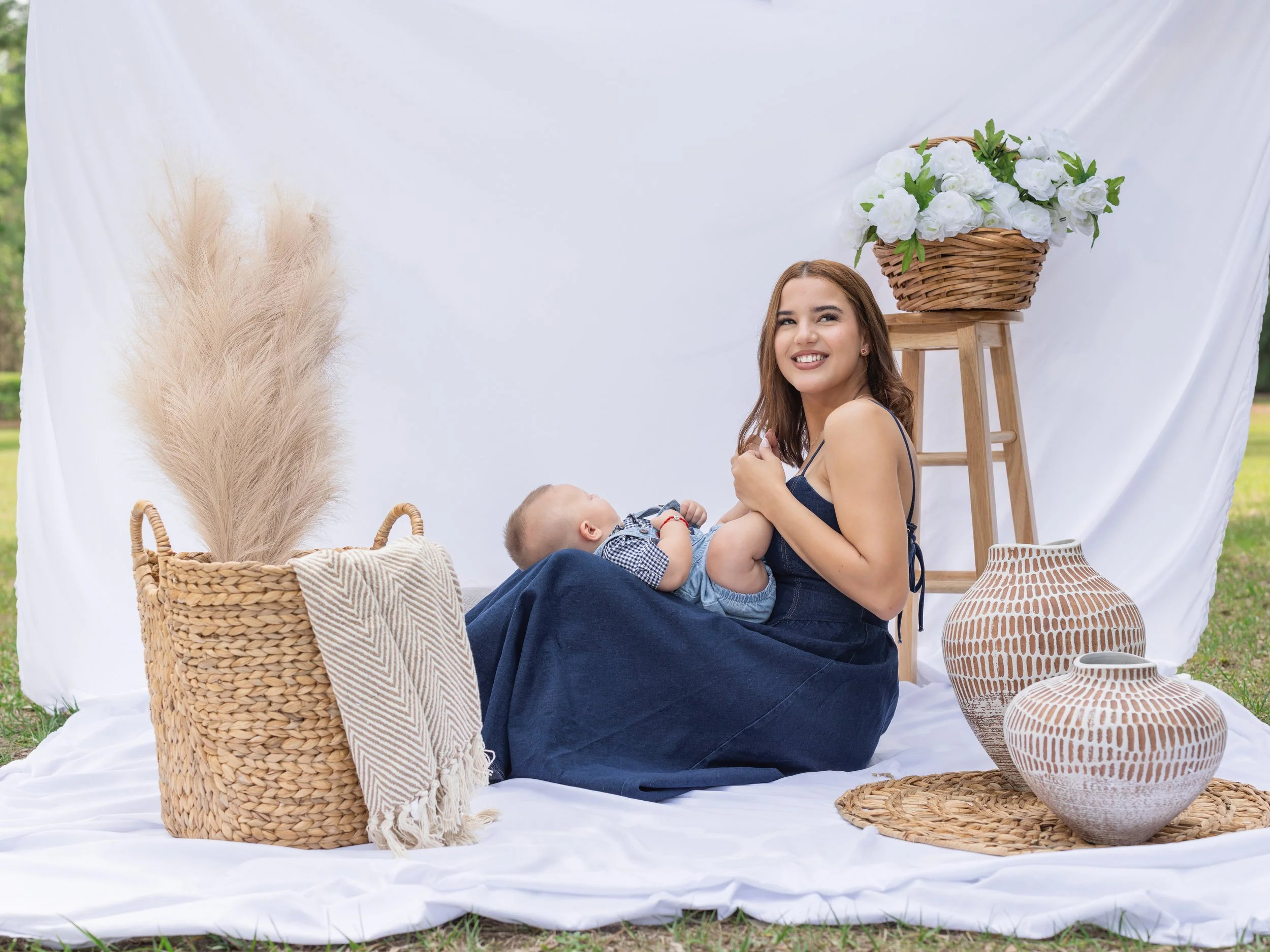 Young woman sitting on a white cloth beside a baby, with wicker baskets, a wooden step ladder, potted plants, and decorative vases around her, outdoors with a white backdrop.