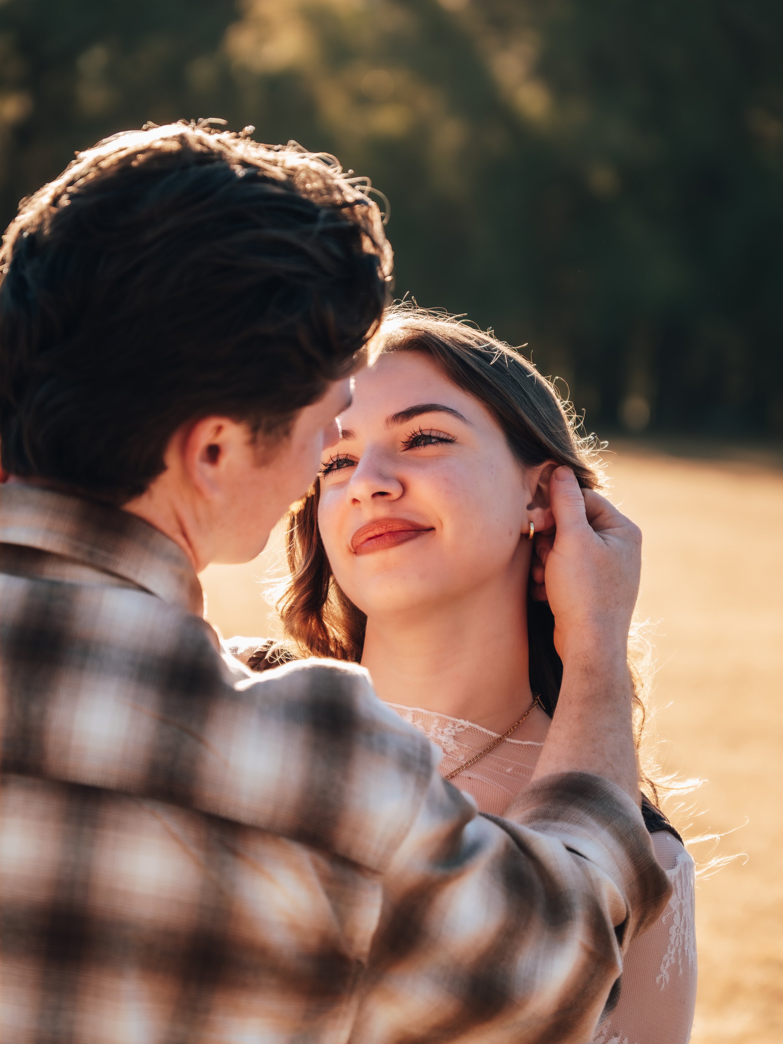 A man gently touches a woman's face as they share an intimate moment outdoors during sunset, with trees in the background ar SHalom Park in florida