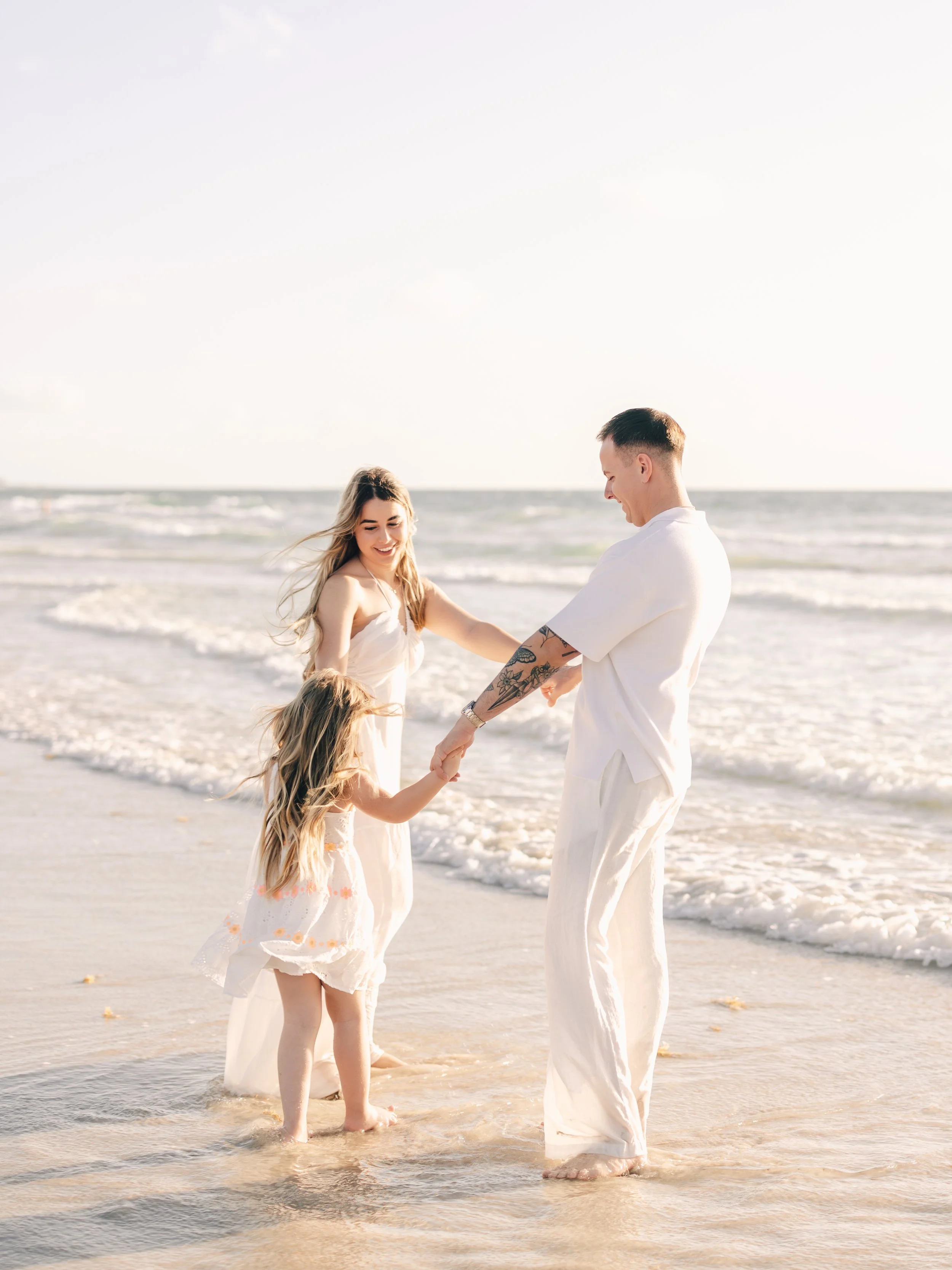 A family of four, including an adult man, a woman, and two young girls, enjoying a walk on the beach with the ocean in the background. They are holding hands and smiling.