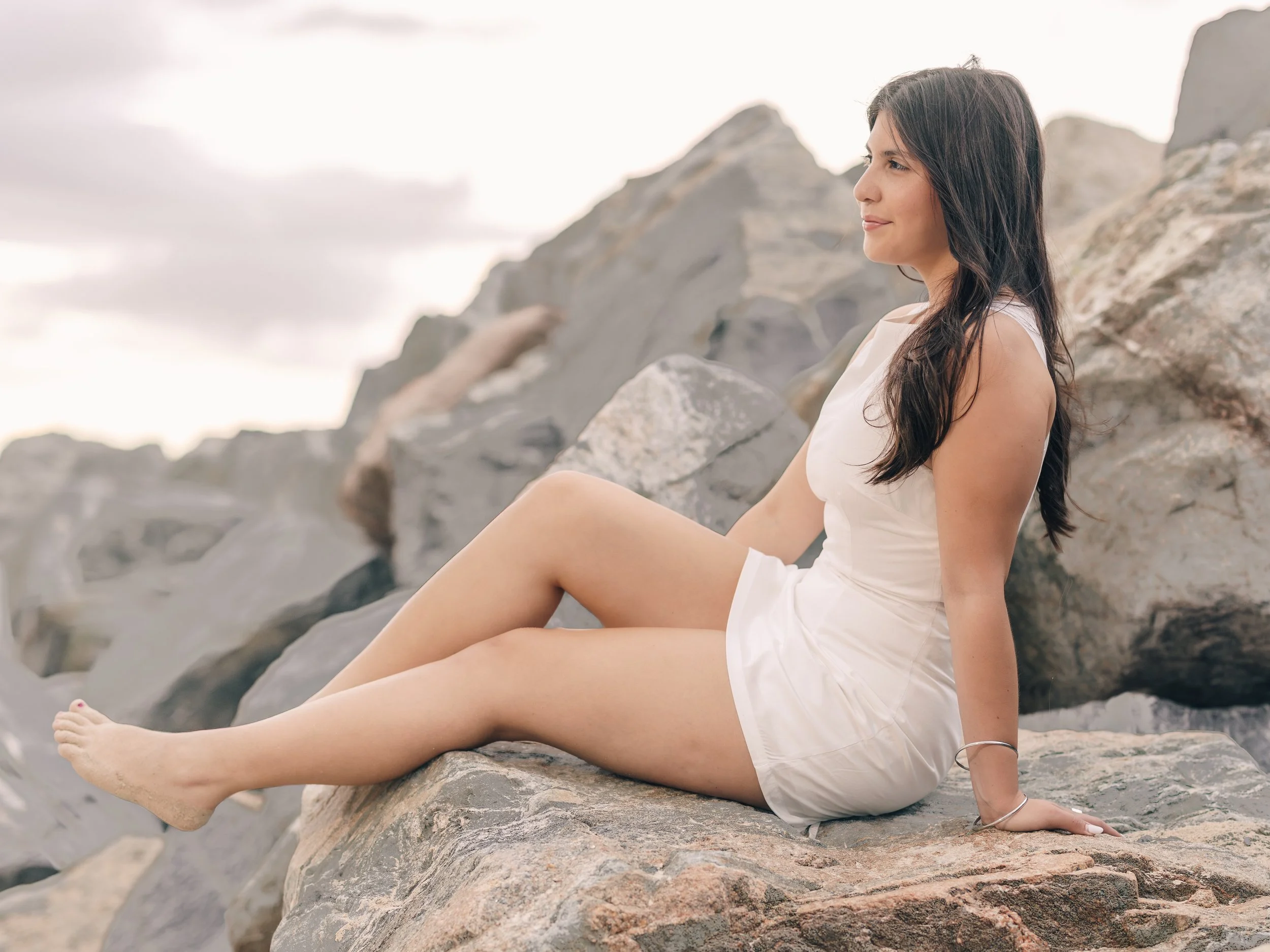 A woman with dark hair sitting on rocks at the beach, smiling slightly, wearing a white sleeveless dress, with a cloudy sky in the background.