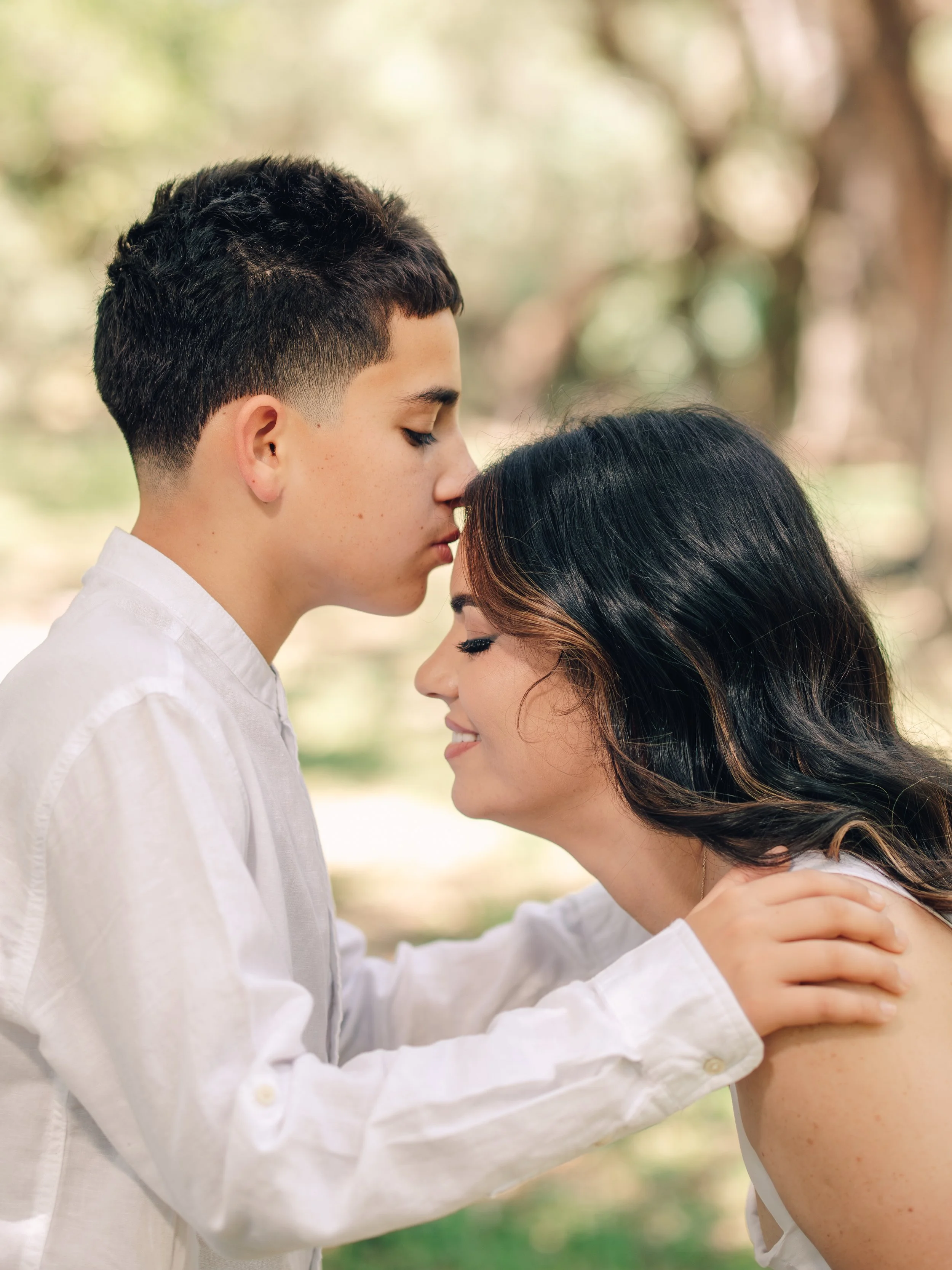 A young man and woman touching foreheads and smiling outdoors with blurred trees in the background.