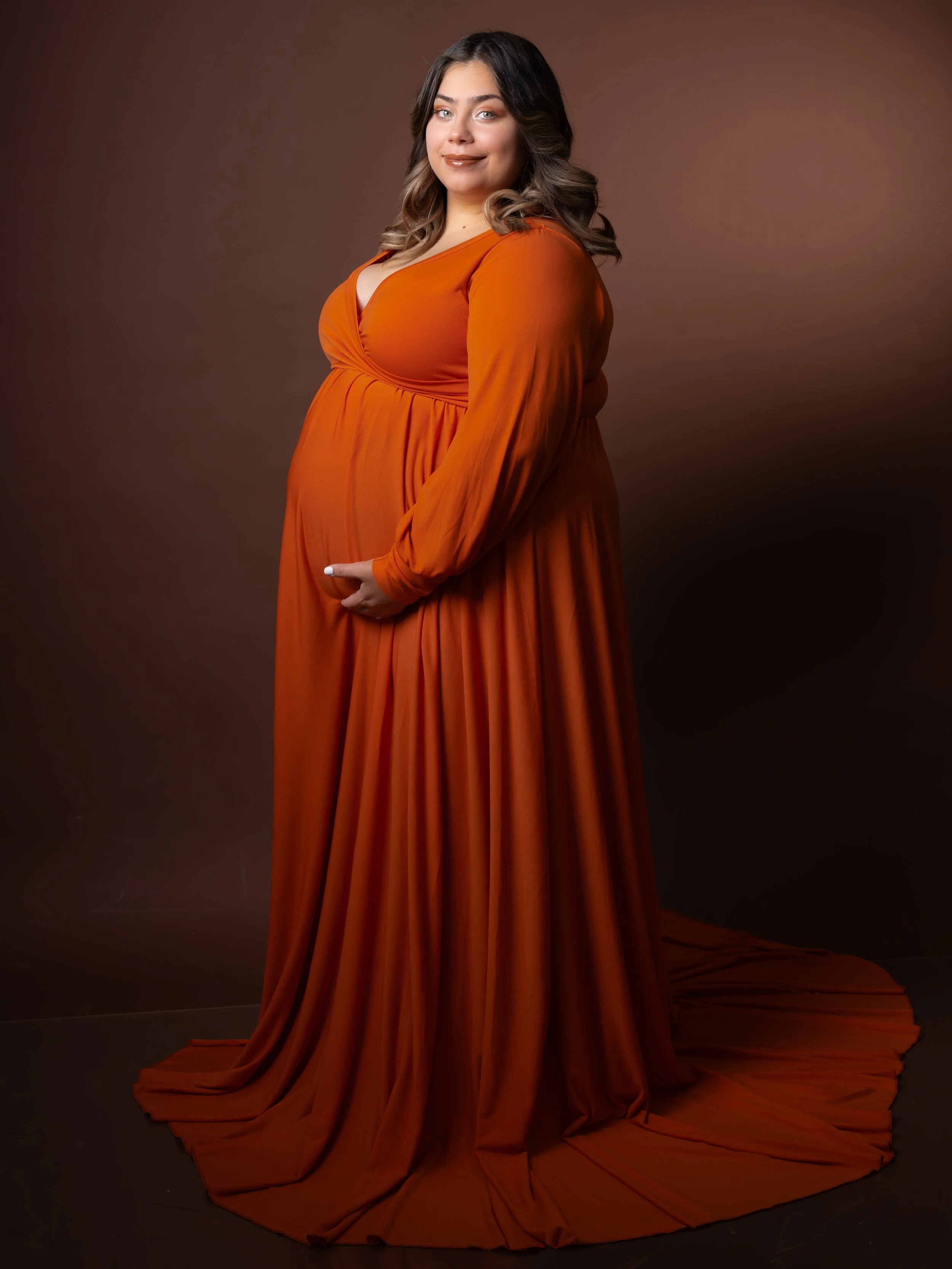 A pregnant woman with shoulder-length brown hair and light skin wearing an orange gown against a brown gradient background.