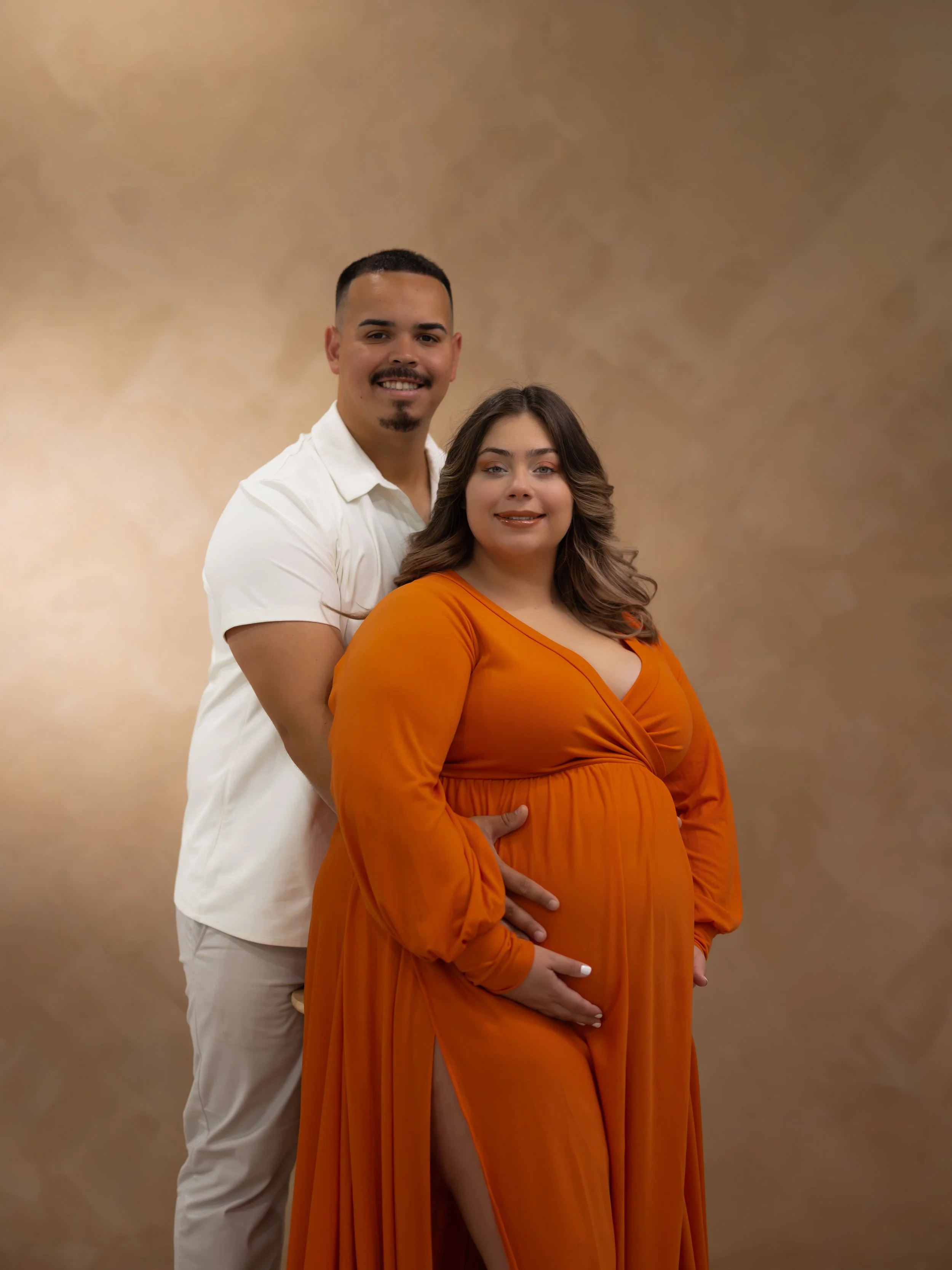 A smiling couple standing close together, the woman is pregnant and wearing an orange dress, while the man is wearing a white shirt and light-colored pants. They are holding the woman's belly and are positioned against a plain, beige background.
