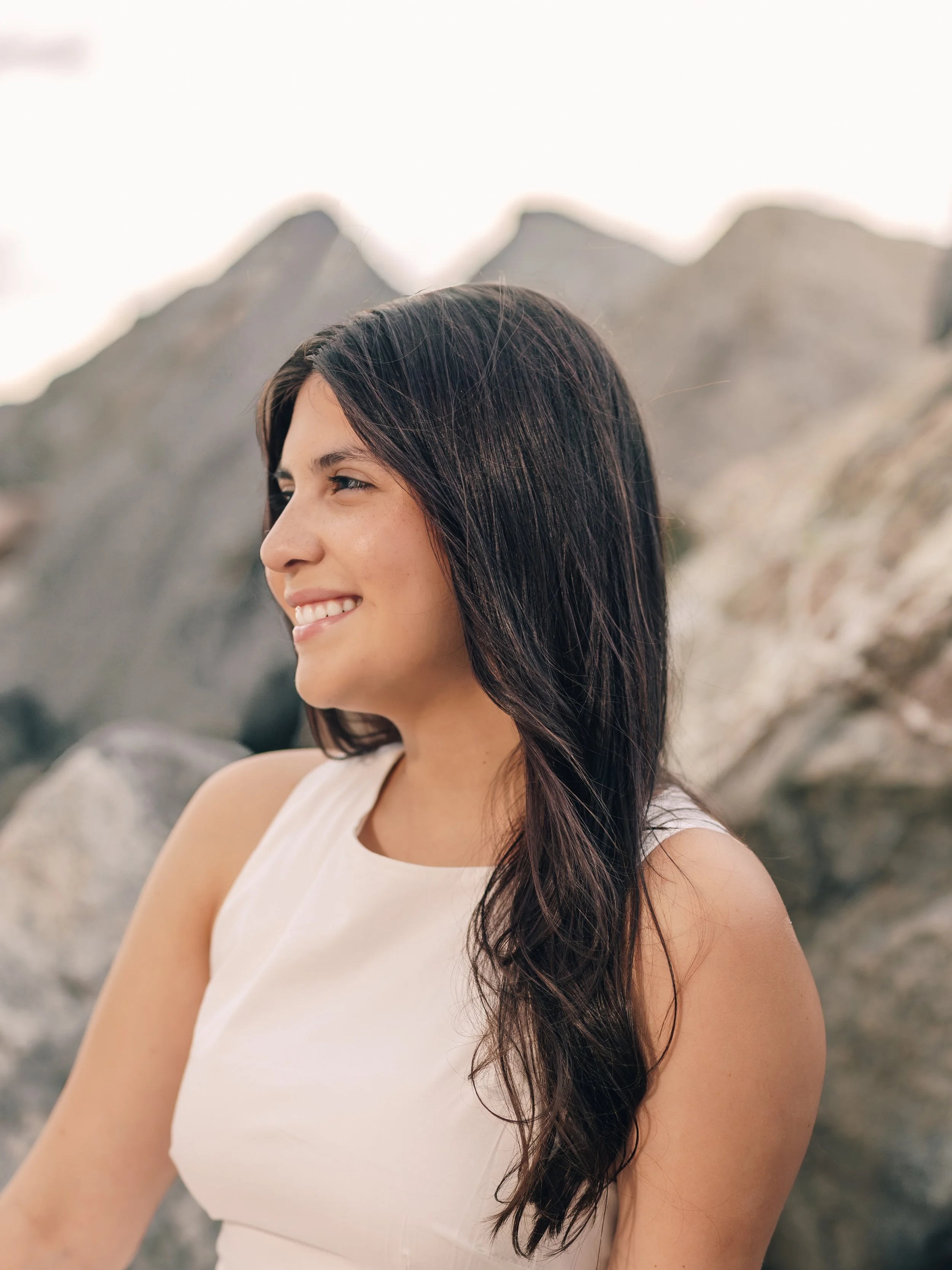 A woman with long dark hair smiling outdoors, with mountains in the background.