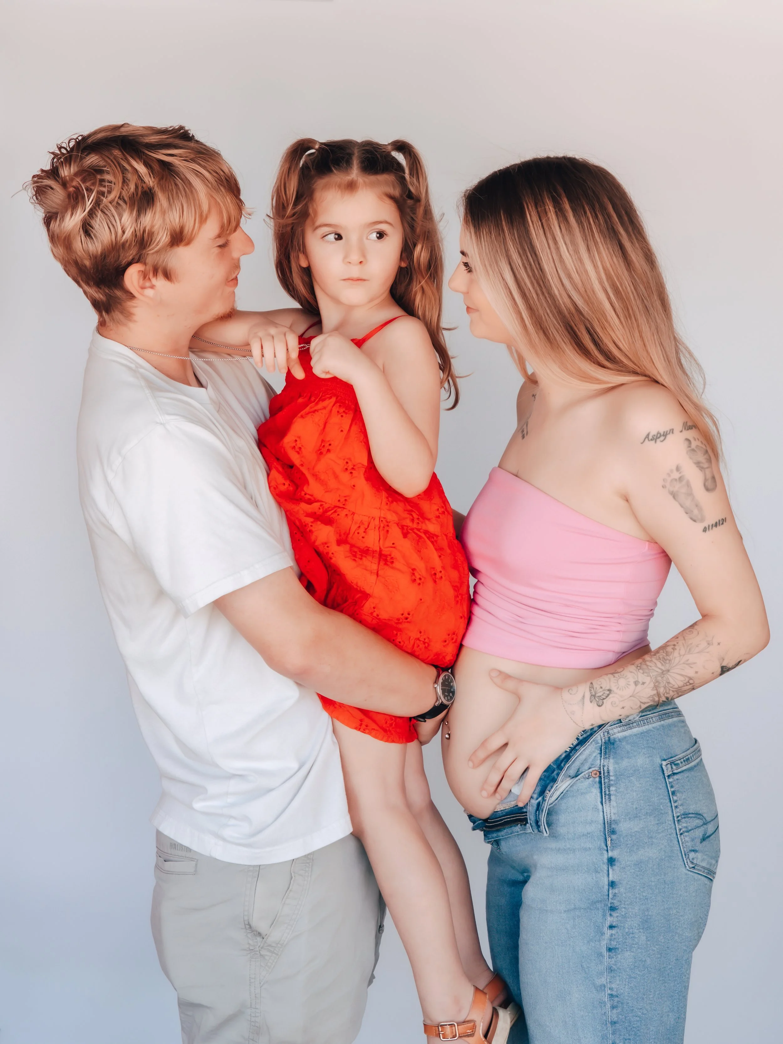 A young girl in a red dress being held by a man, with a woman in a pink top and jeans looking at them, all standing against a plain light gray background.