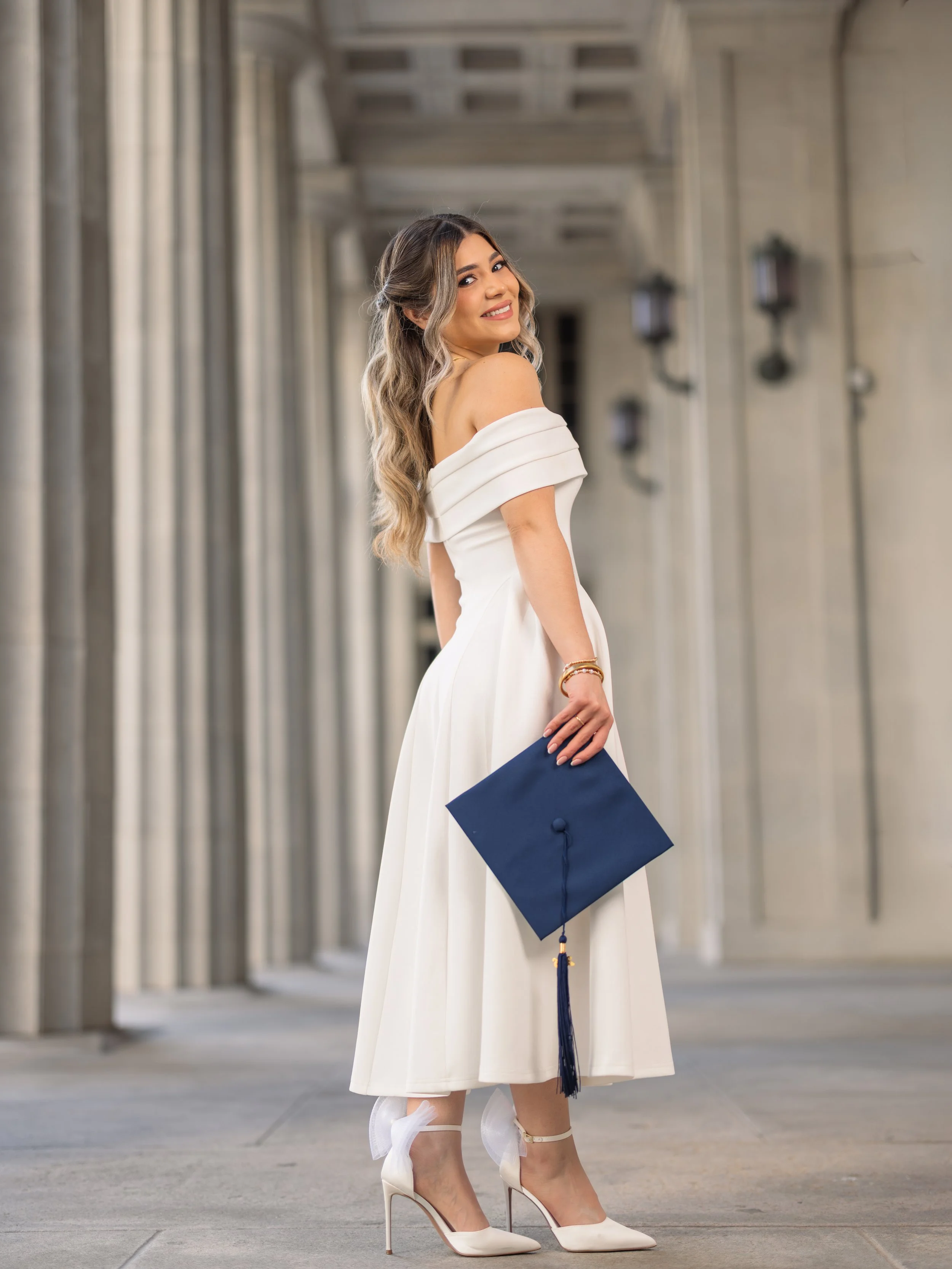 A young woman dressed in a white off-the-shoulder dress with white high heels, holding a navy blue graduation cap with a tassel, standing outside in front of a building with large columns.
