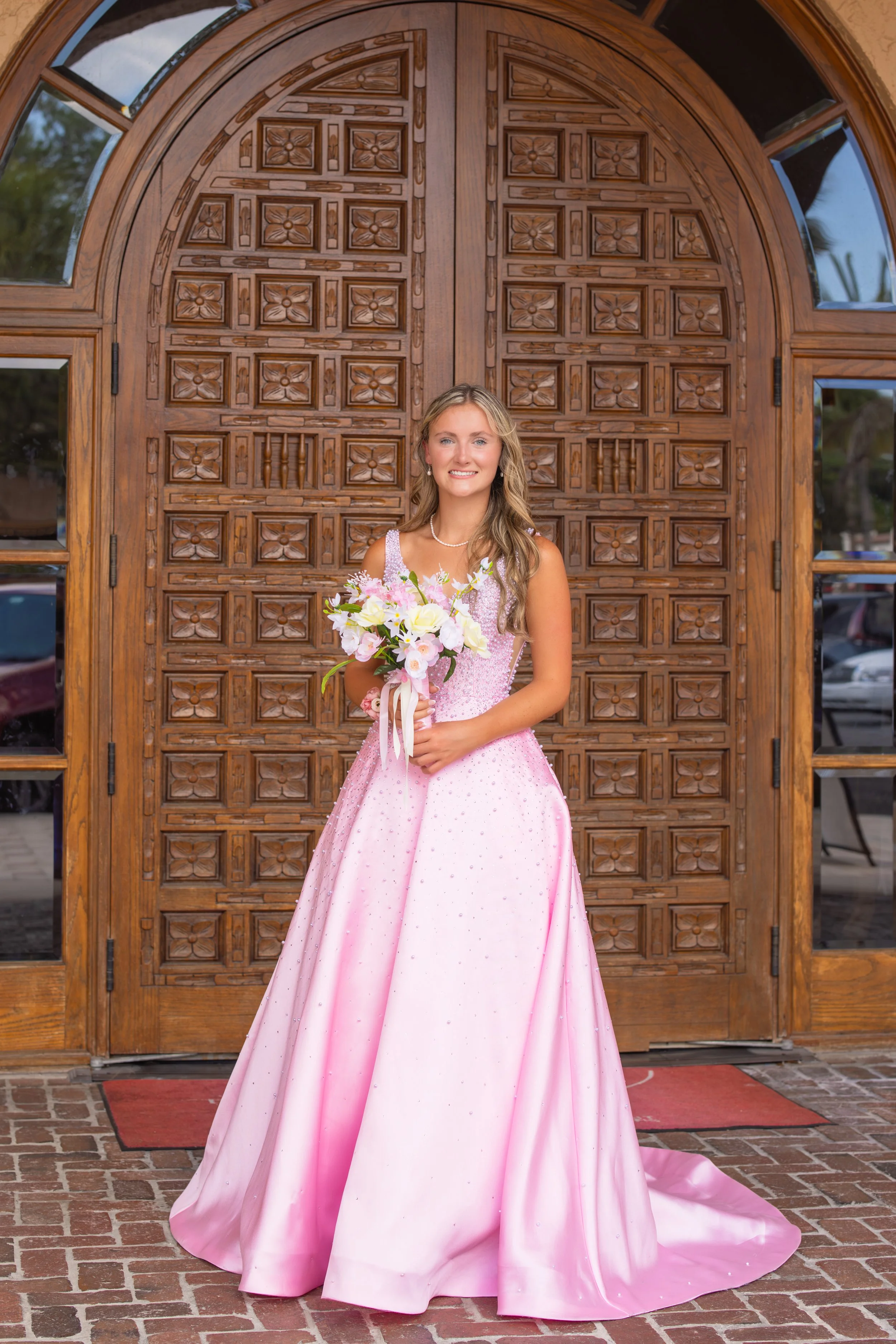 A young woman in a pink ball gown standing in front of an ornate wooden door holding a bouquet of flowers.