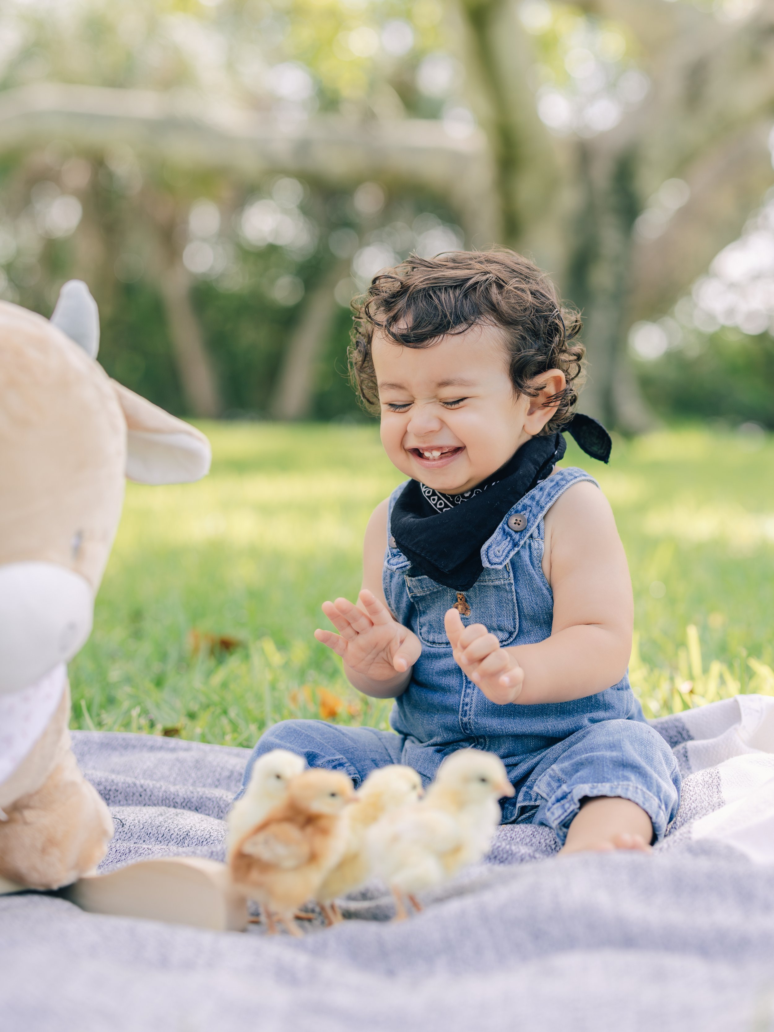 A young child with curly hair, wearing a black bandana and denim overalls, sitting on a blanket outdoors surrounded by ducklings, smiling with eyes closed.
