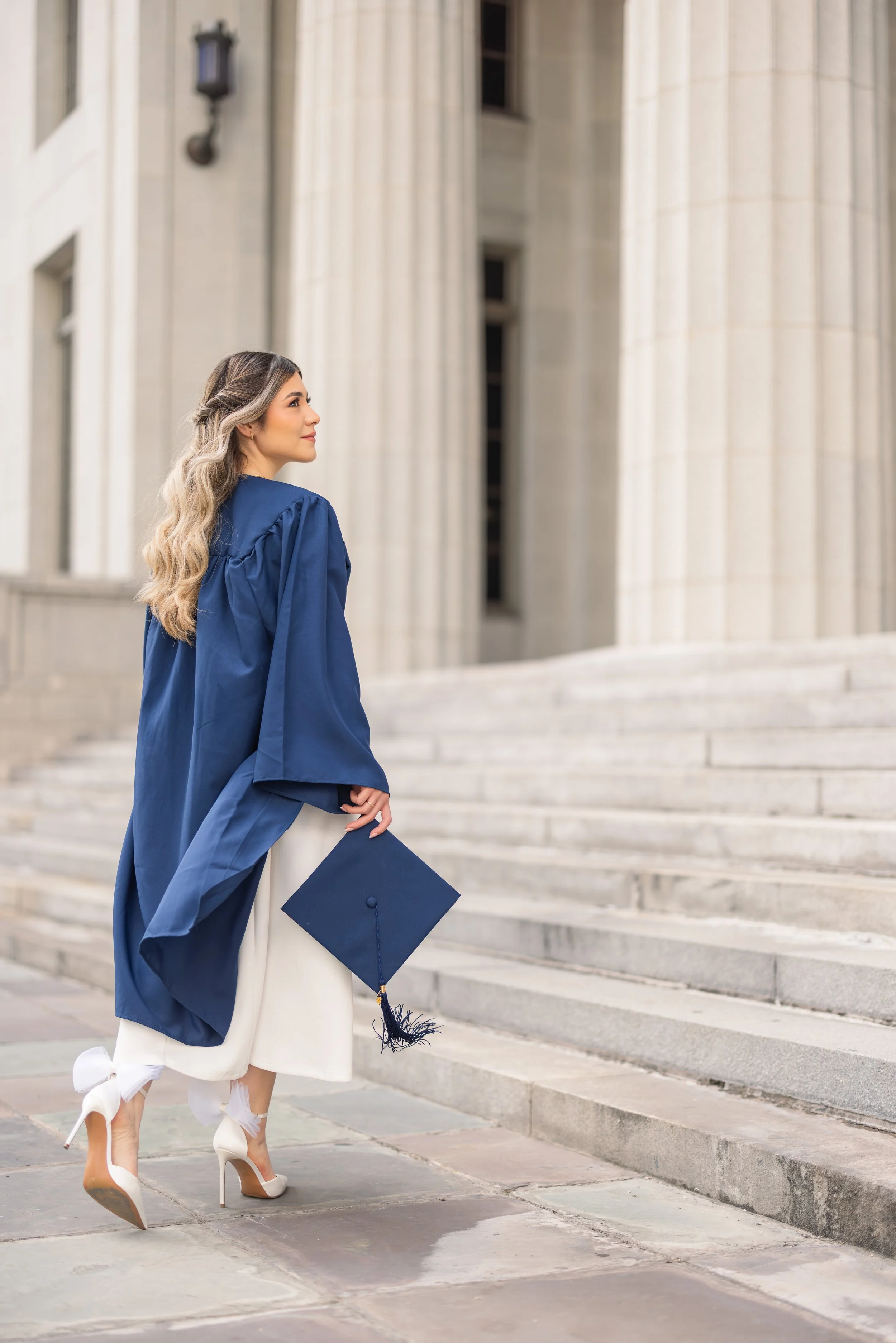 A young woman in a graduation gown holding a diploma walking up steps outside a stone building with columns.