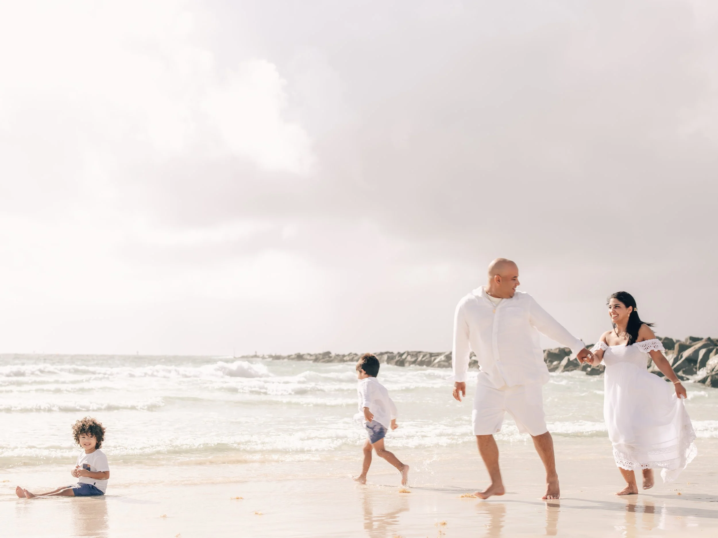 A family of four dressed in white, walking and playing on a sandy beach with ocean waves, rocks, and a cloudy sky in the background.