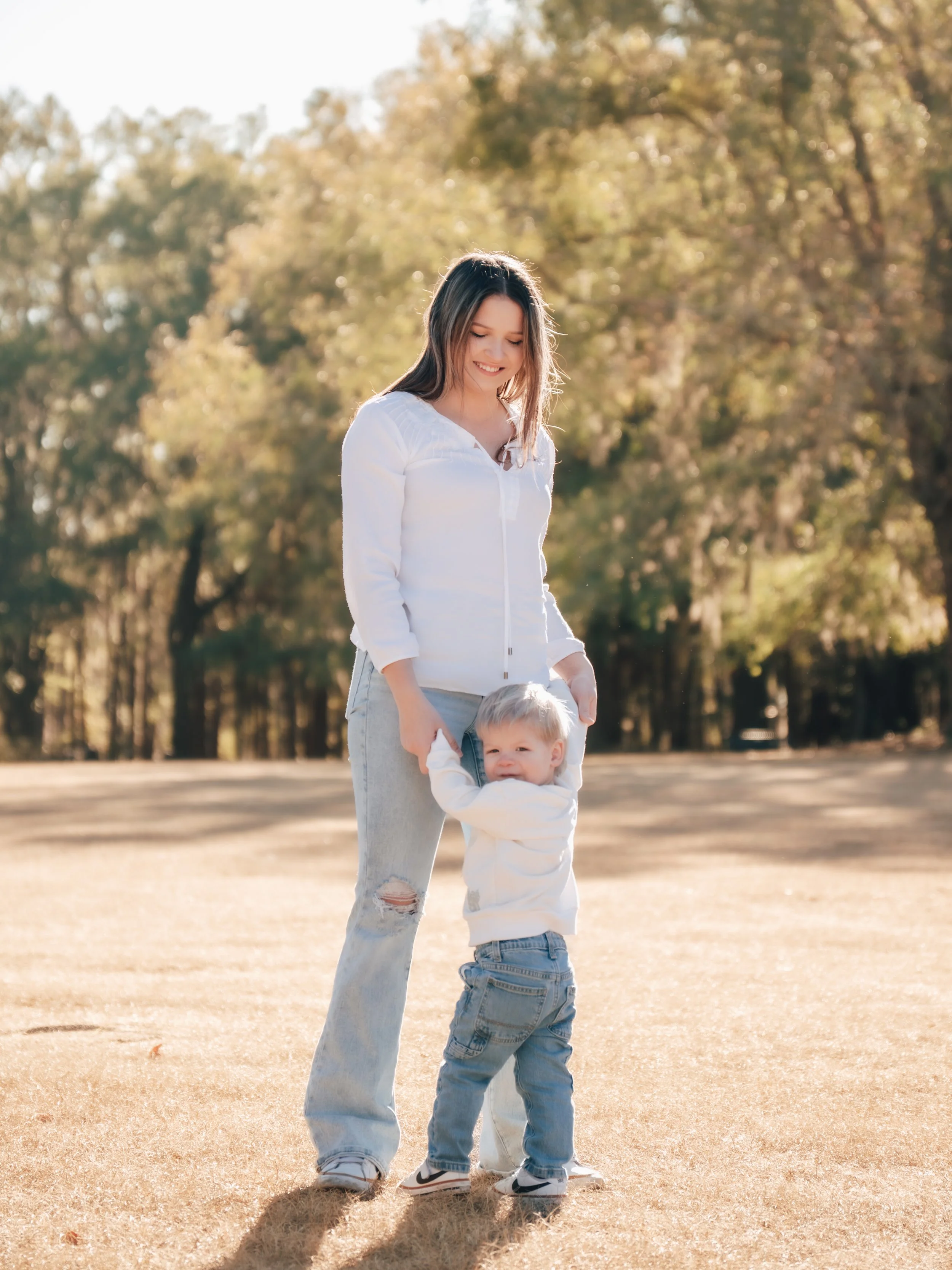 A woman and a young boy playing outside in a park with trees in the background. The woman is smiling and holding the boy's hands as he stands and faces the camera. The boy appears to be smiling. they are in florida