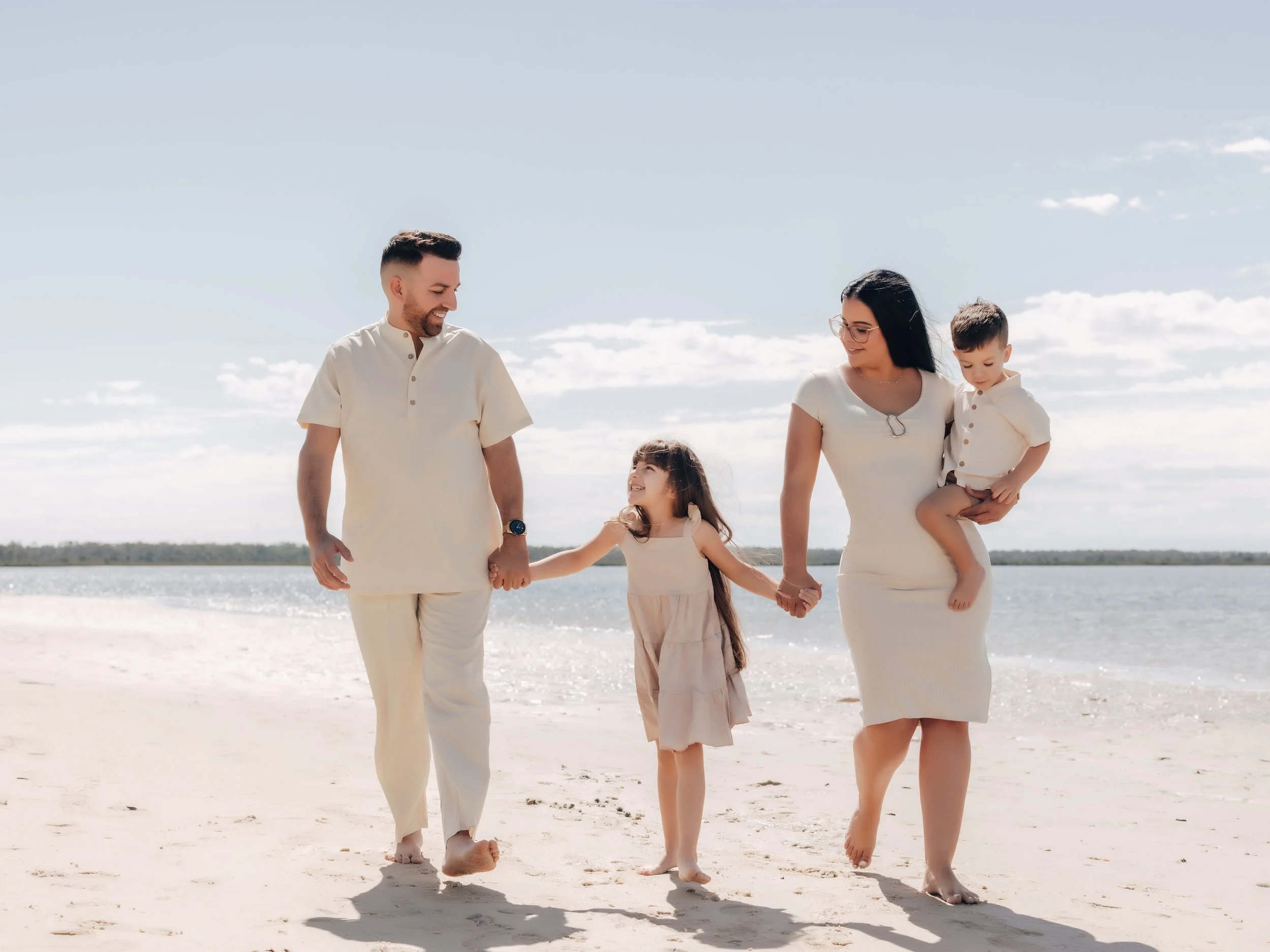 A family of four walking on the beach holding hands, with the ocean and sky in the background.