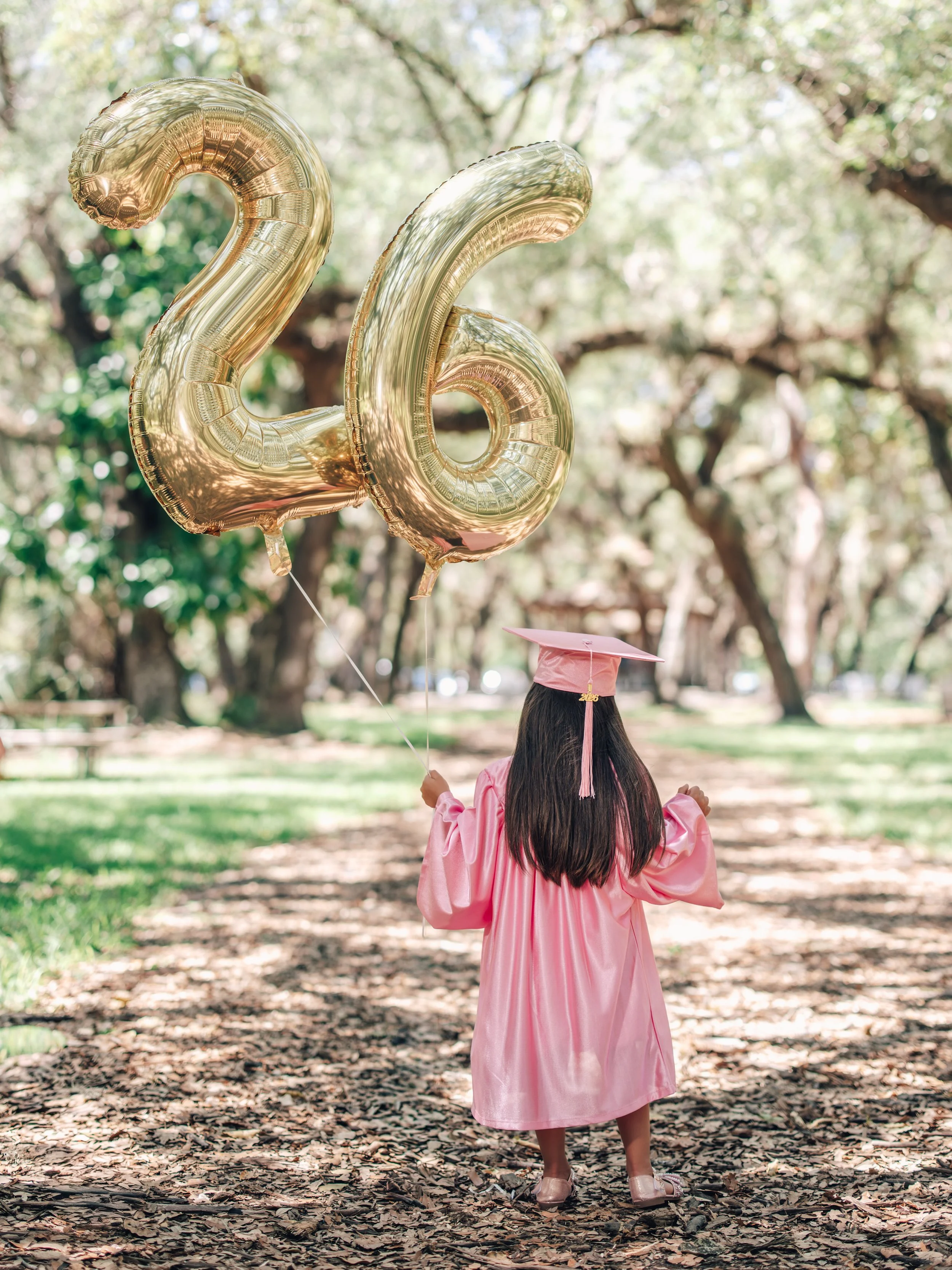 A young girl dressed in a pink graduation gown and cap is holding two large gold foil balloons shaped as the numbers 2 and 6, standing outdoors on a path covered with fallen leaves and surrounded by trees.