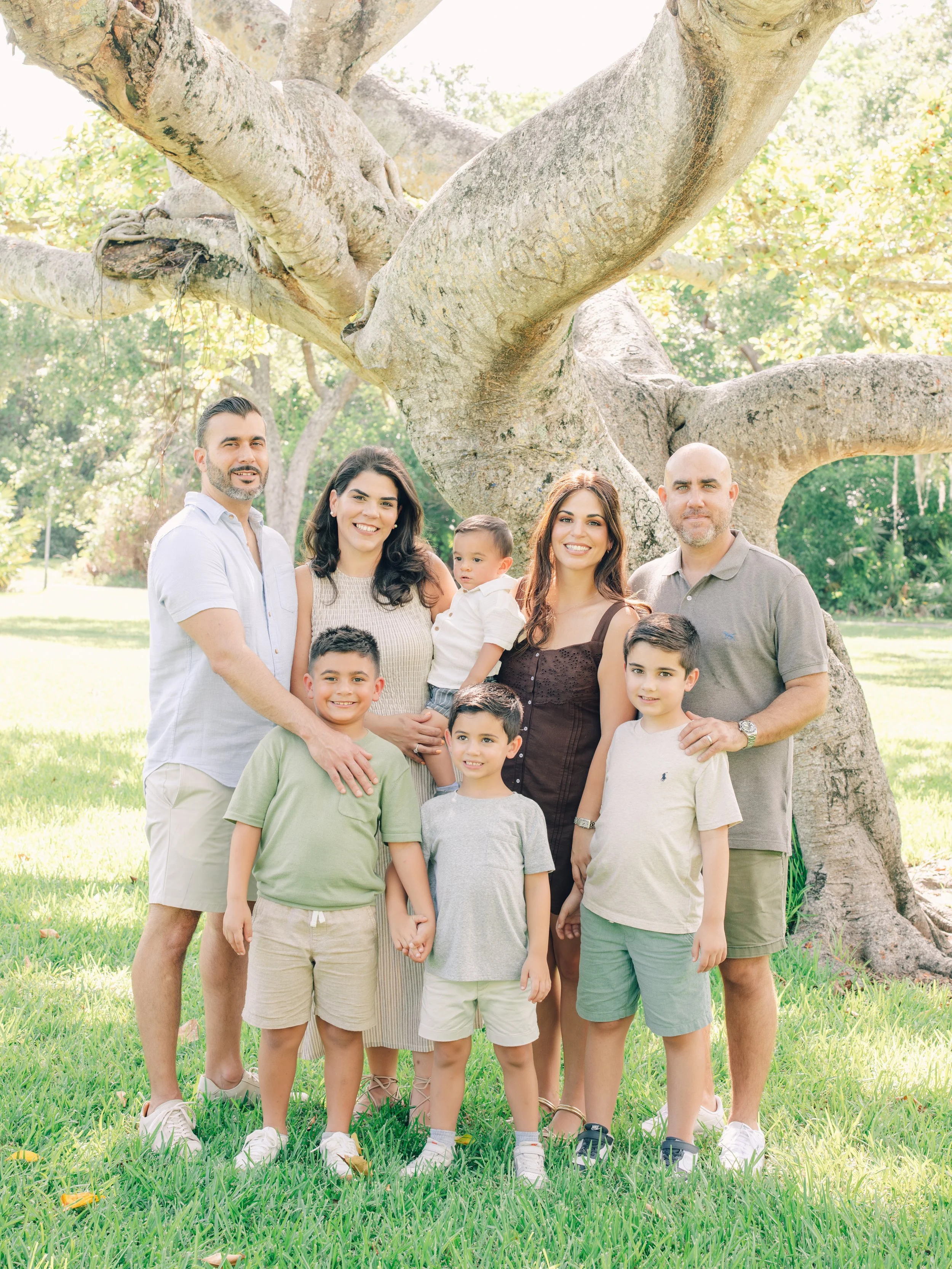 Group of nine people, including adults and children, standing outdoors under a large tree with sprawling branches and green leaves. They are smiling and posing for the photo on a grassy area.