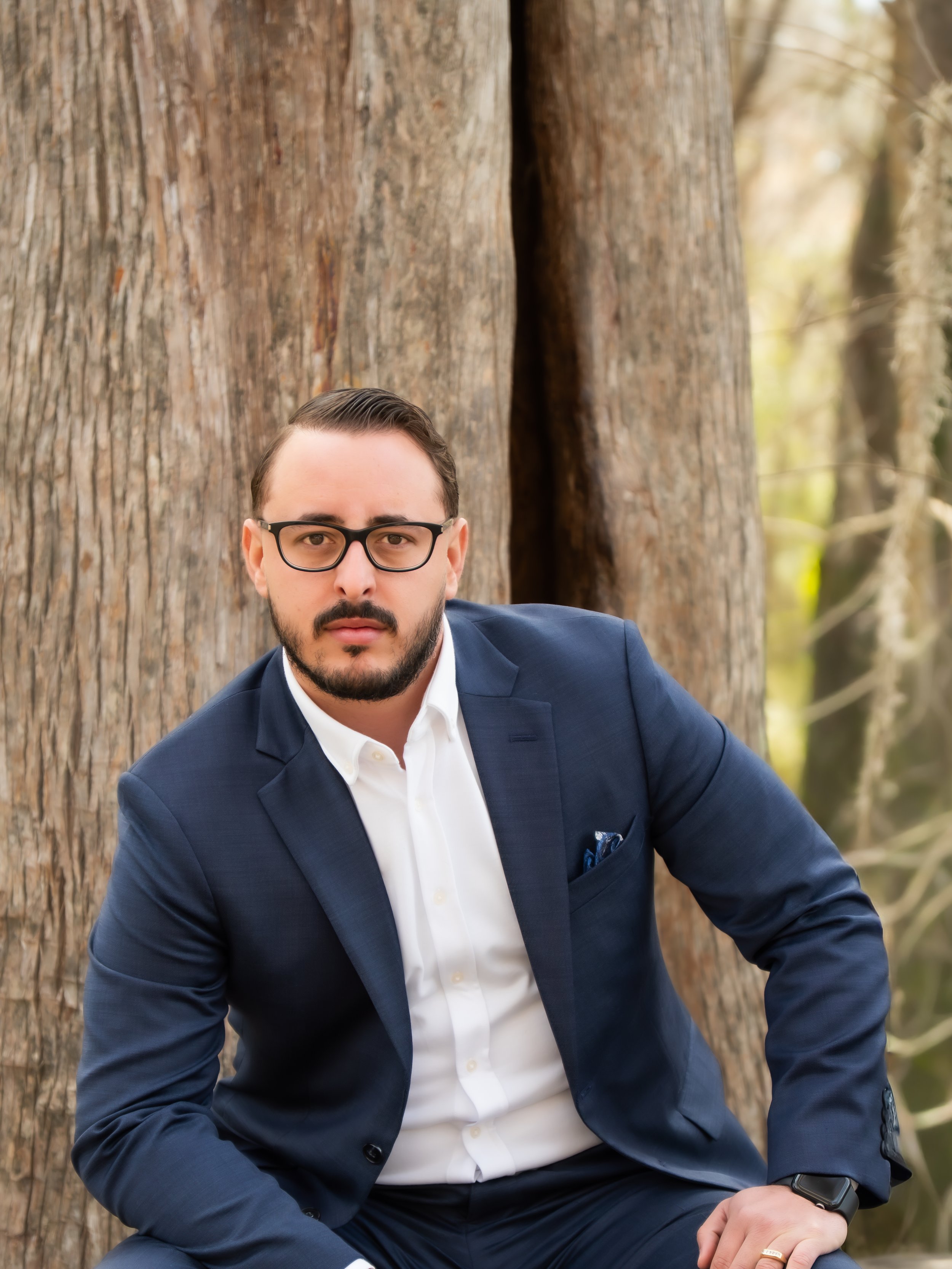 A man in a navy suit and white shirt sitting outdoors near trees with bark, looking at the camera.
